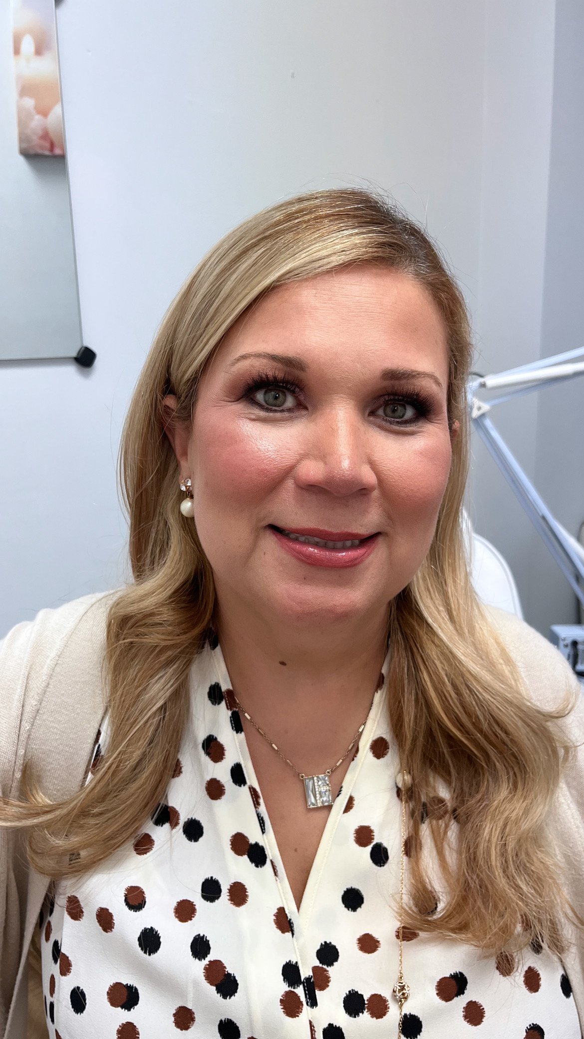 A smiling woman with long blonde hair, blue eyes, wearing pearl earrings, a necklace with a large square pendant, and a cream-colored blazer over a polka-dot blouse, sitting in an office or medical setting.