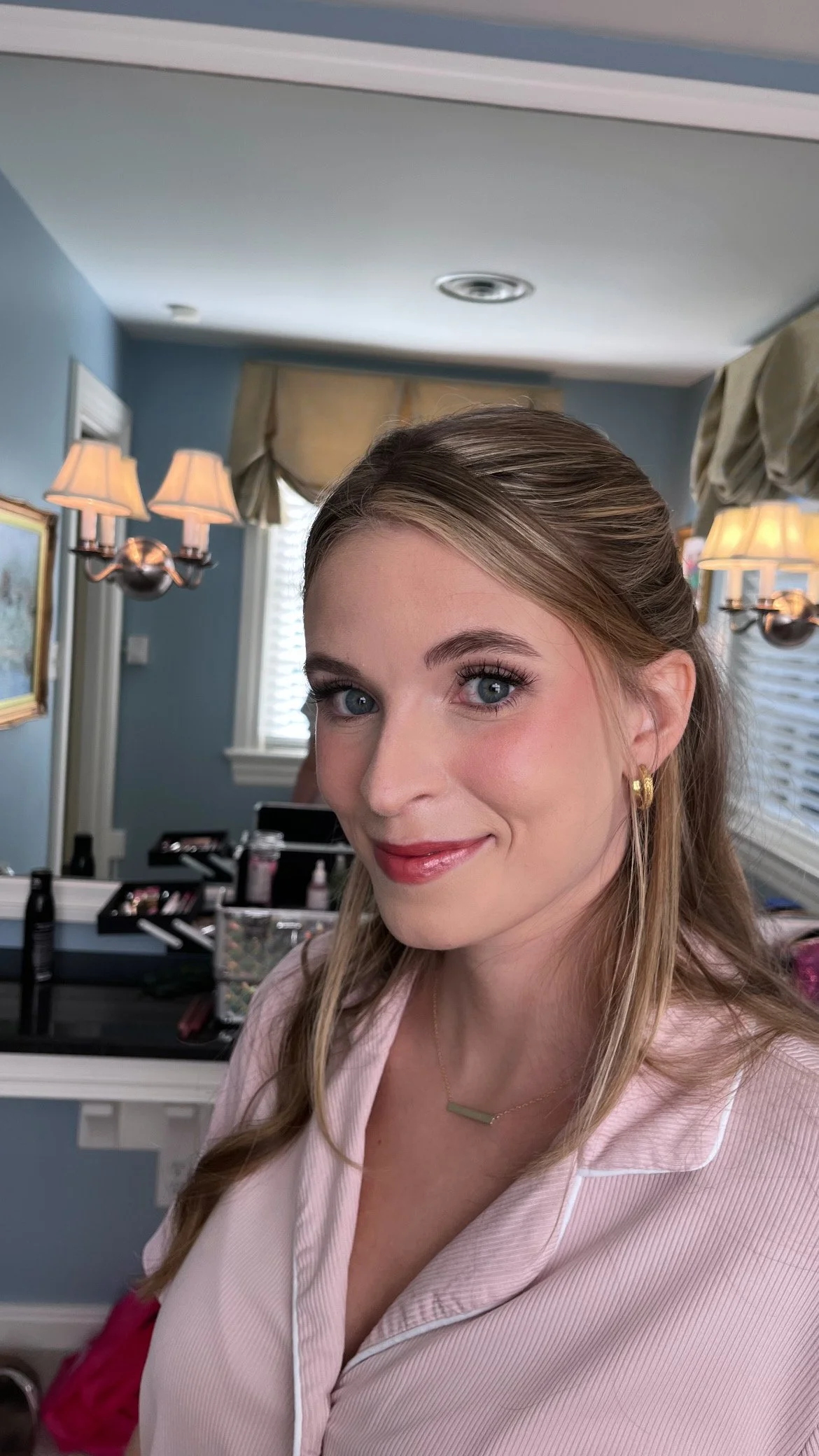 A woman with blonde hair styled in loose waves, wearing a light pink top with white piping, gold hoop earrings, and a gold bar necklace, smiling at the camera in a room with blue walls, window blinds, curtains, and a vanity mirror with makeup and bea