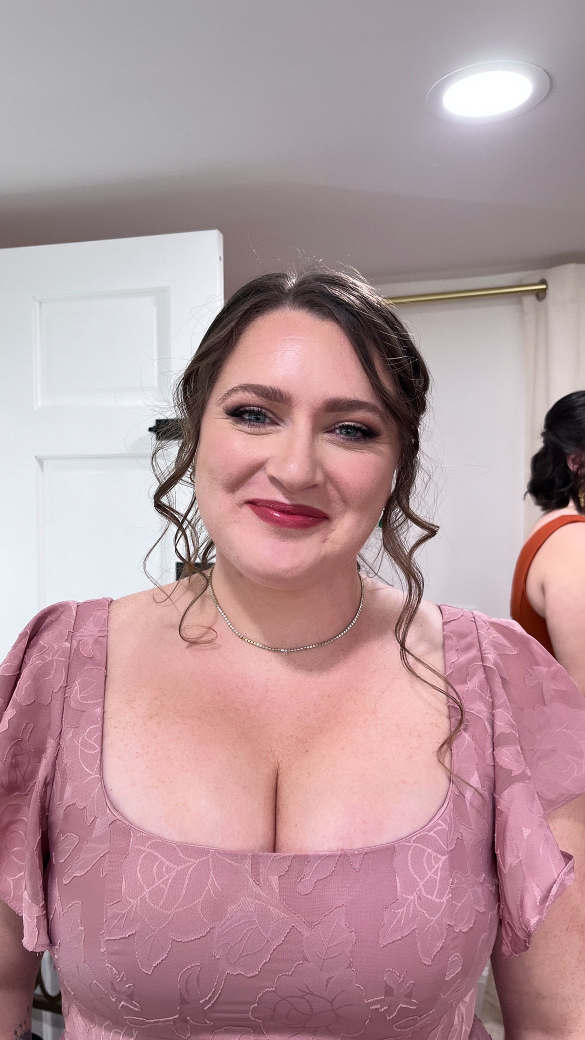 A woman with wavy brown hair wearing makeup and a pink lace dress with a square neckline.