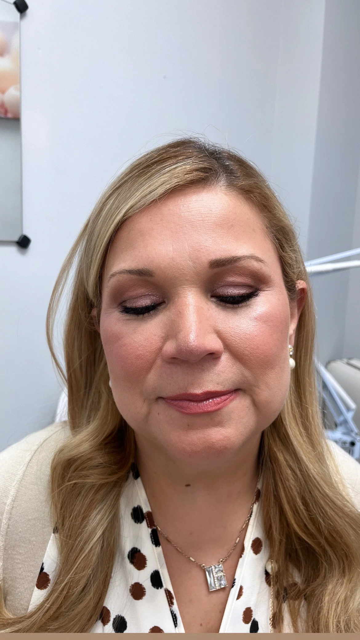 Close-up of a woman with blonde hair, wearing makeup, a polka dot blouse, a necklace, and earrings, in a clinical or office setting.