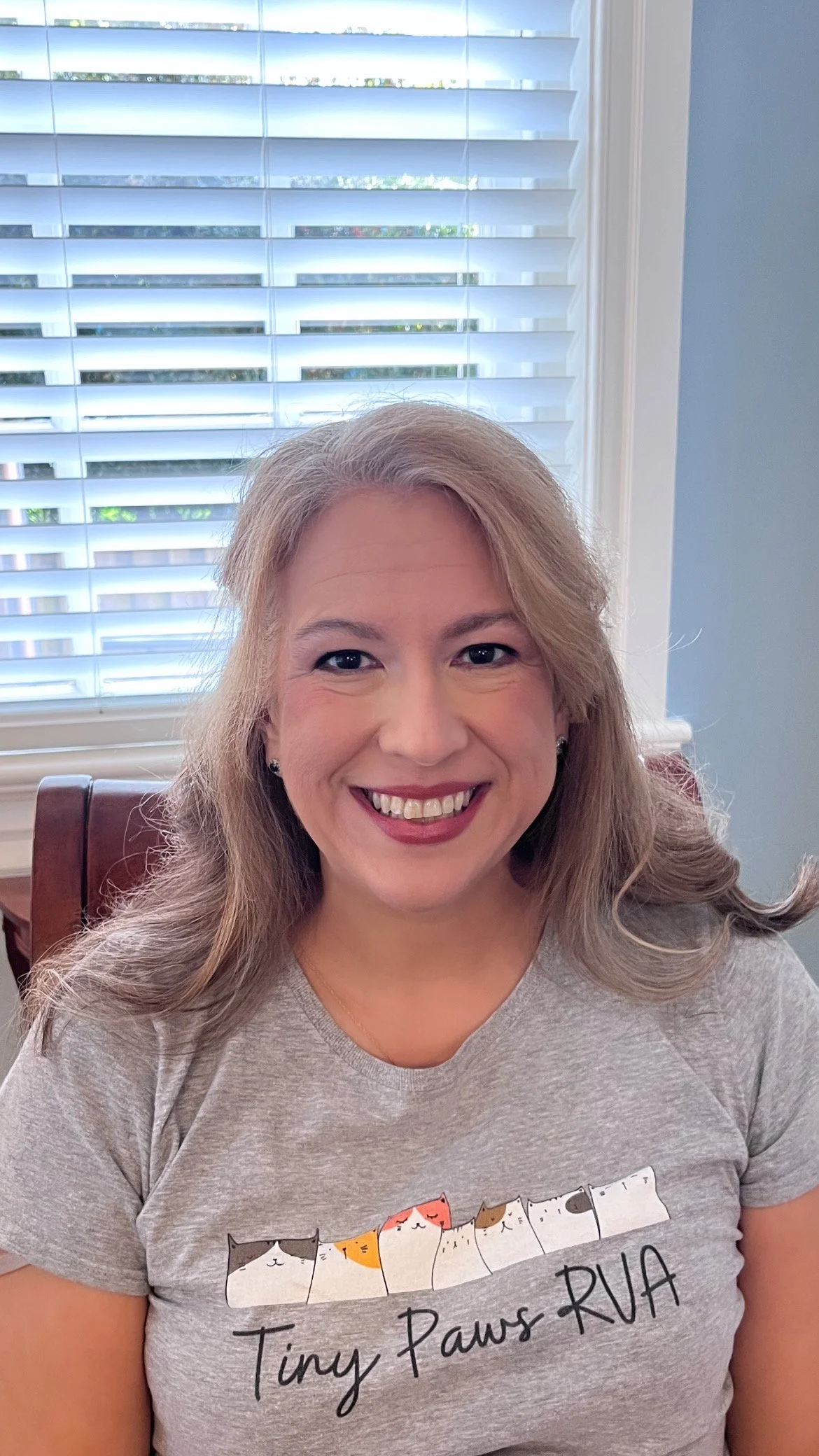 A woman smiling, sitting in front of a window with white blinds, wearing a gray T-shirt with cartoon cats and the words 'Tiny Paws RUA' on it.