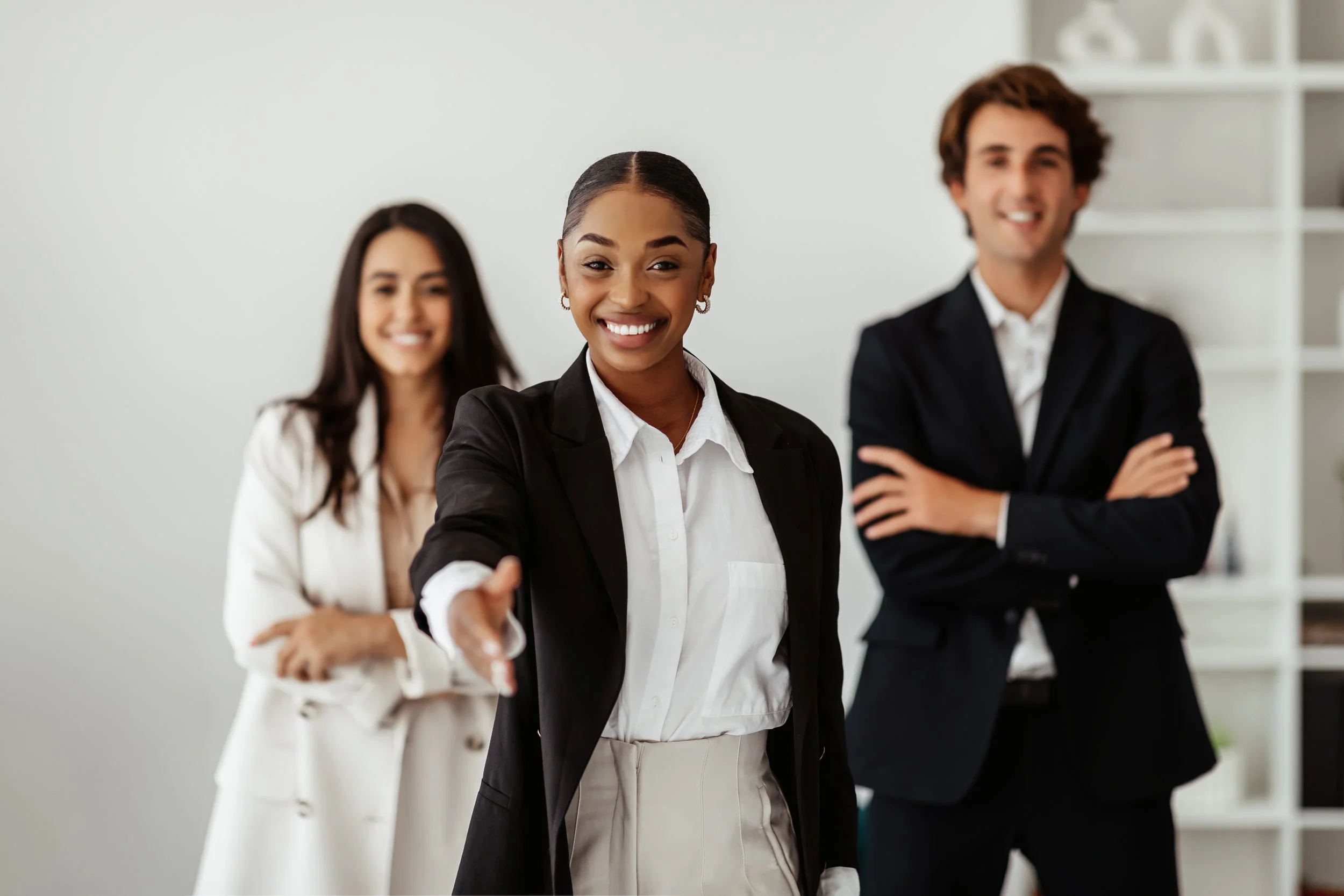 Three business professionals in formal attire smiling, with the woman in the front extending her hand for a handshake in an office setting.