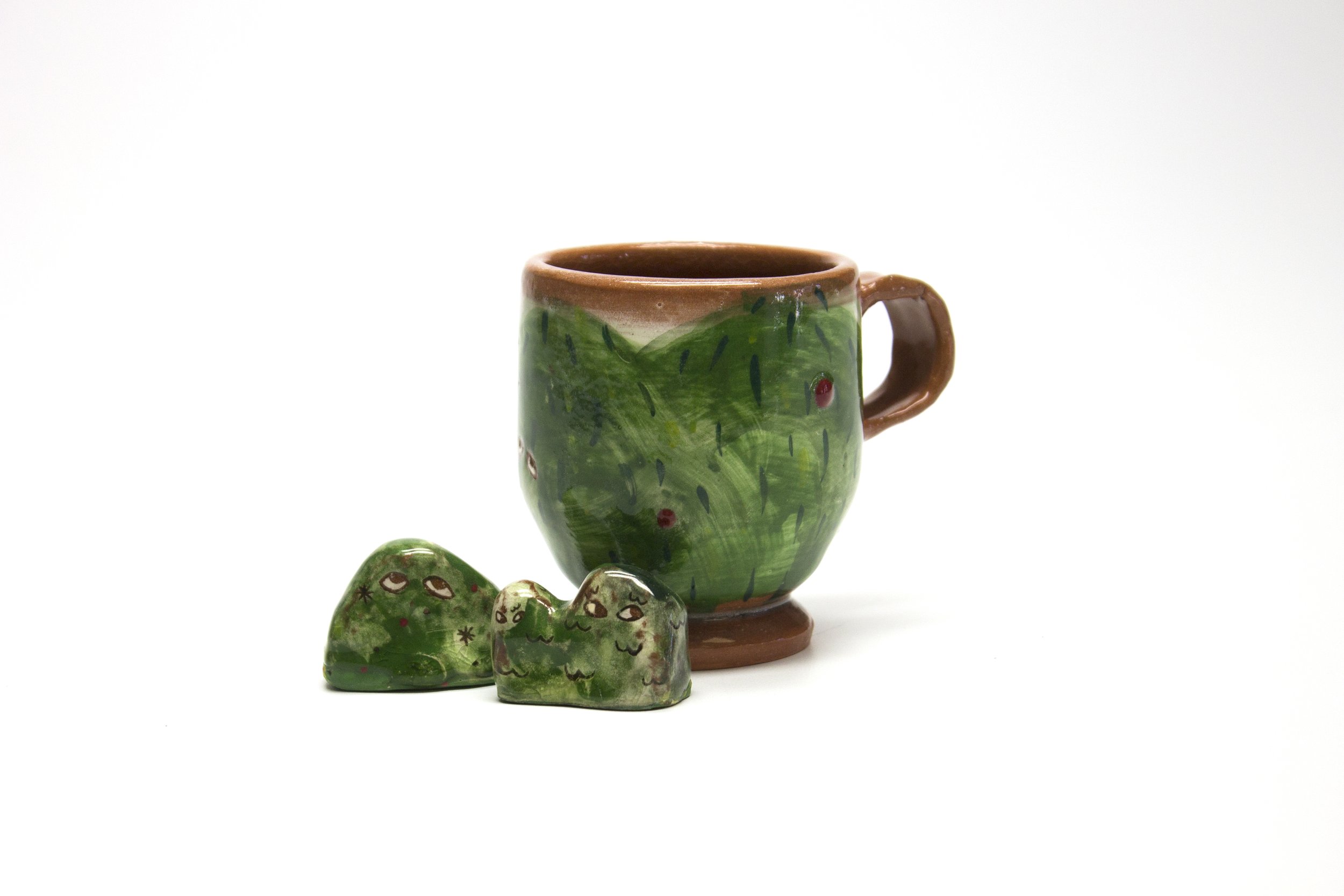 Hand-painted ceramic cup with a green leafy design and two small heart-shaped ceramic objects with similar design, all against a white background.