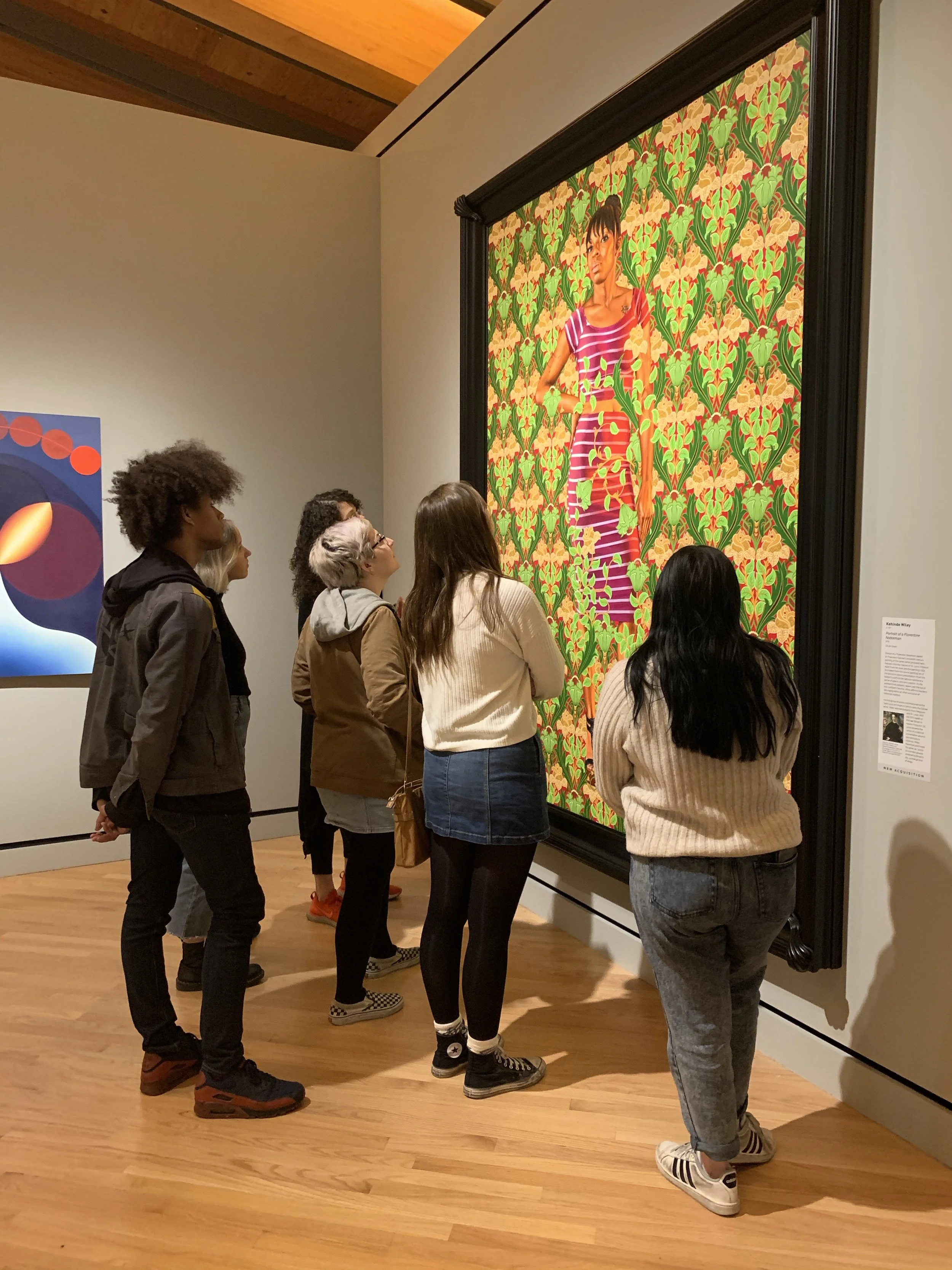 Gallery of people observing a colorful painting of a woman in a striped dress surrounded by floral patterns at an art museum.