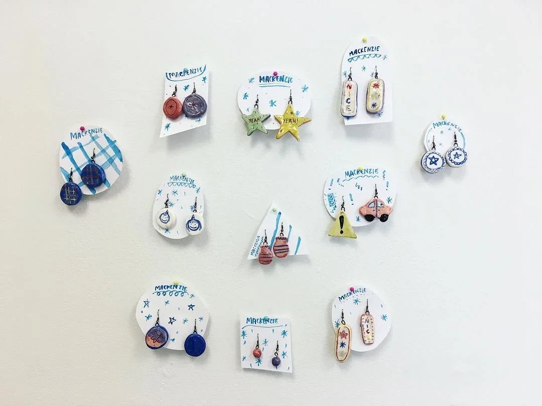 Display of various handmade earrings on a white wall, each pair attached to a decorated card labeled "MACKENZIE." The earrings feature different shapes, colors, and themes, including stars, circles, animals, and holiday motifs.