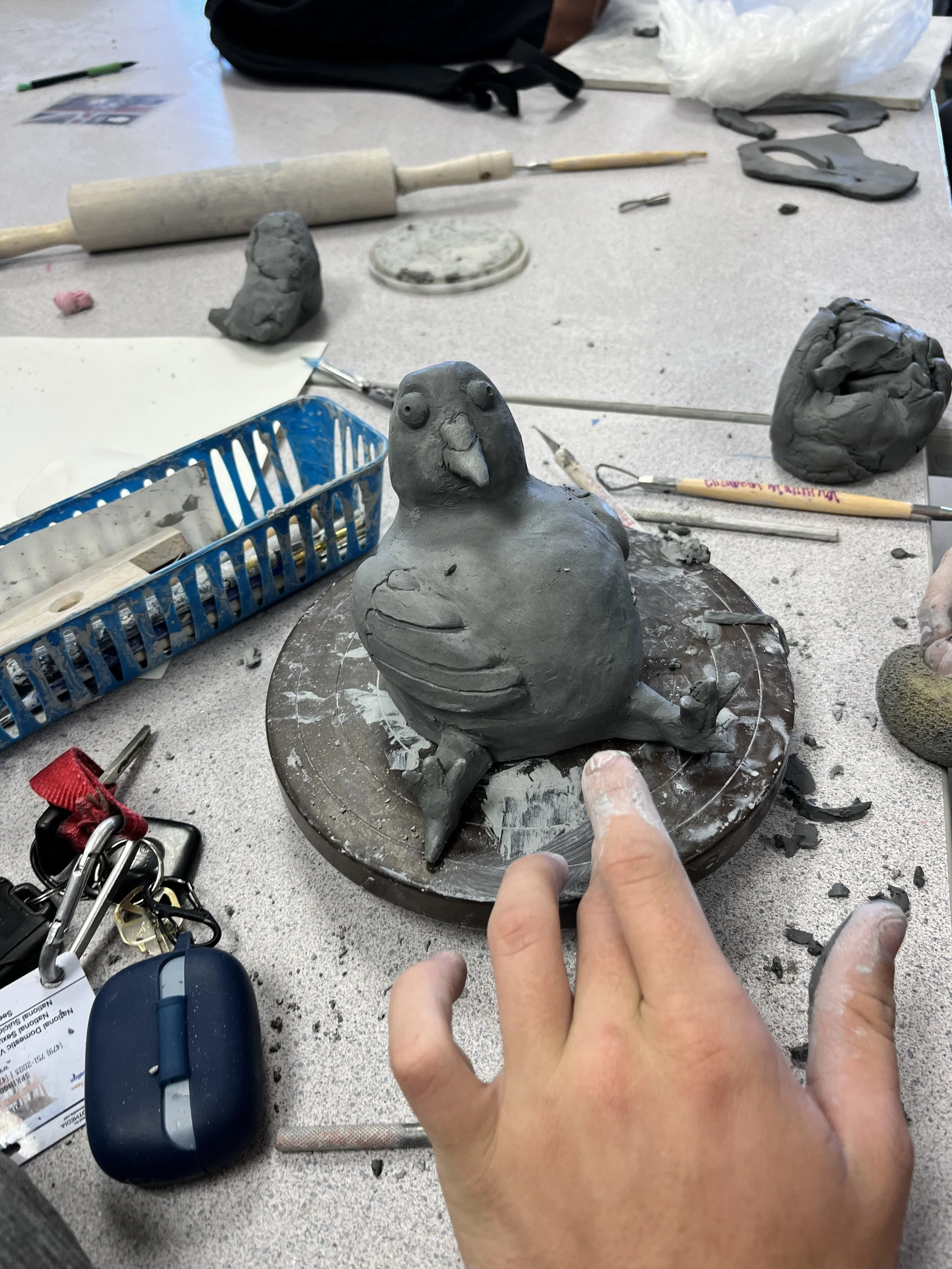 A person working on a clay sculpture of a bird, possibly a pigeon or dove, in a classroom or studio setting. The sculpture is on a turntable, surrounded by sculpting tools, clay pieces, and drafting supplies.