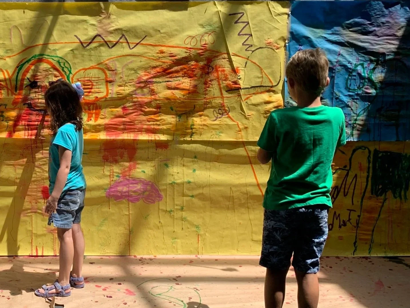 Two children standing in front of a large yellow paper sheet with colorful doodles and drawings, with one girl on the left wearing a blue T-shirt and denim shorts, and a boy on the right wearing a green T-shirt and patterned shorts, both observing the artwork.