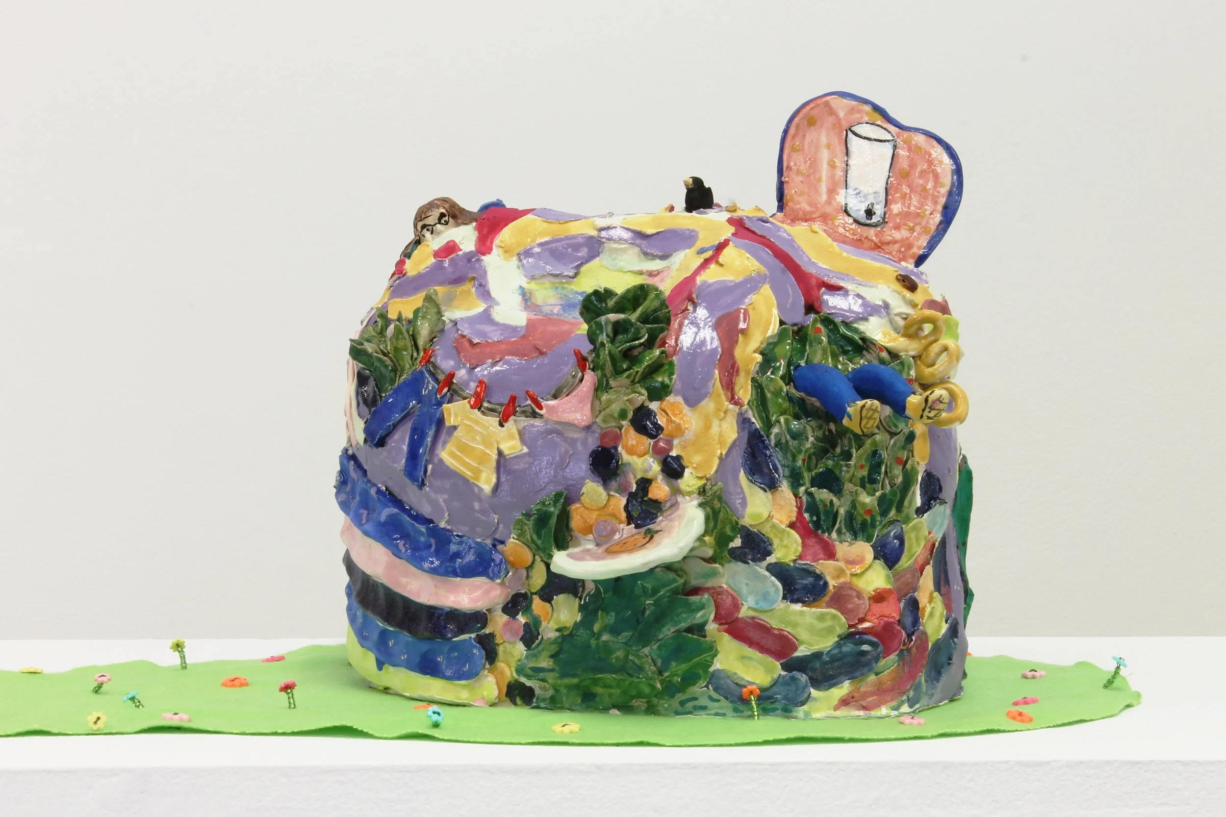Colorful abstract sculpture resembling a house or structure with various painted and textured details, placed on a green base with small decorative elements.