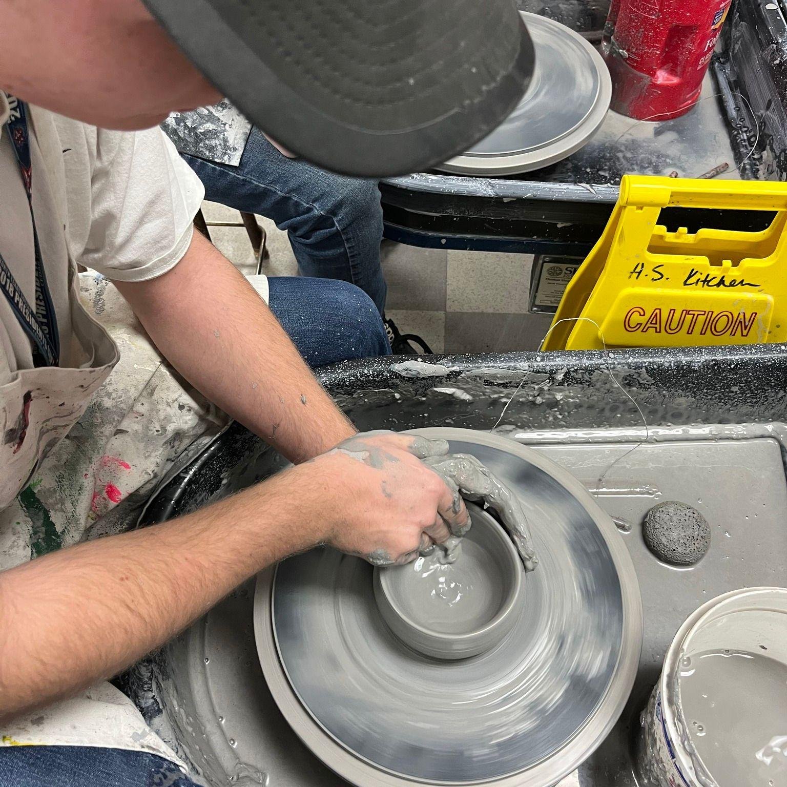 A person is shaping wet clay on a pottery wheel. There is a bucket of clay nearby and a yellow caution sign on the table that says 'H.S. Kitchen'.