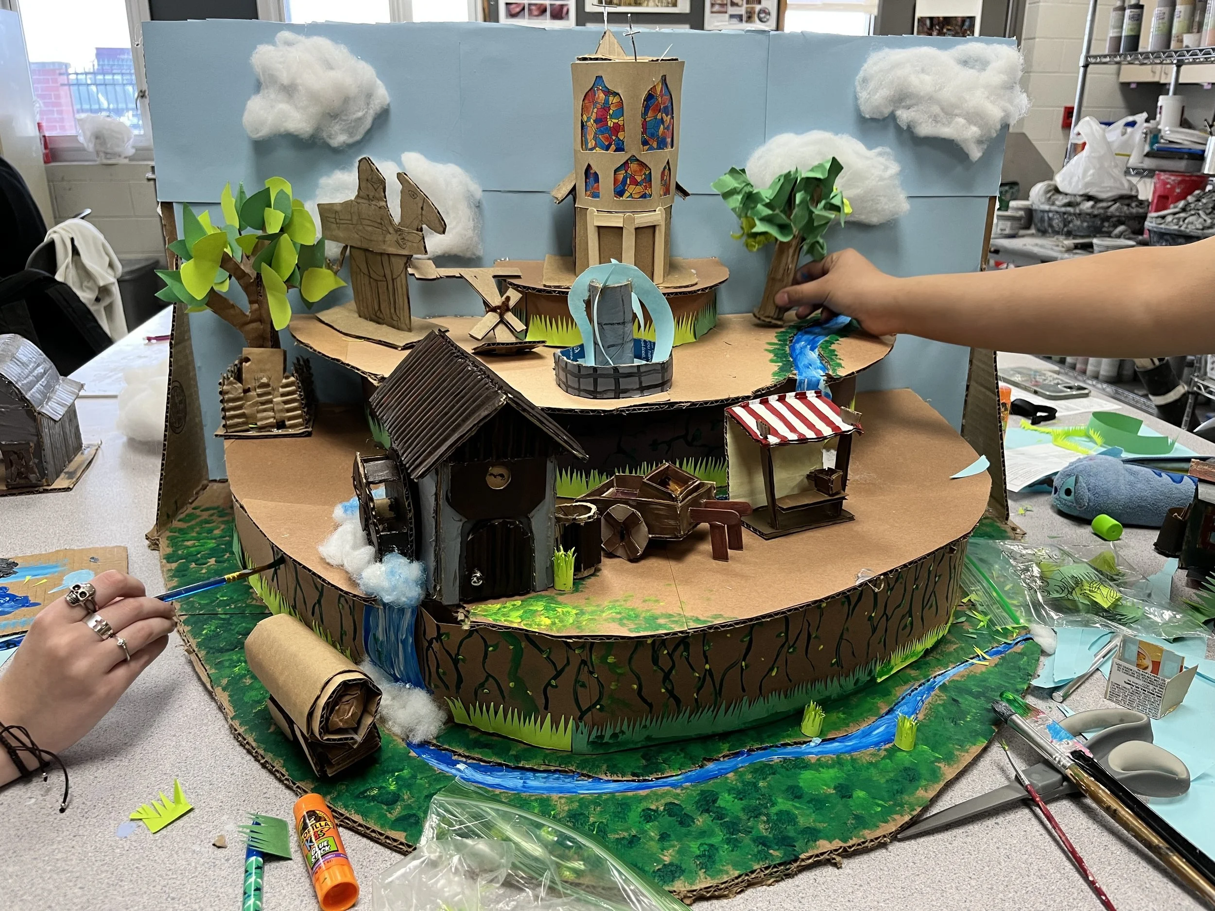 A multi-tiered diorama featuring a castle, trees, a fountain, a river, and various miniature structures made from cardboard, paper, and cotton, with clouds and greenery, in progress on a table.