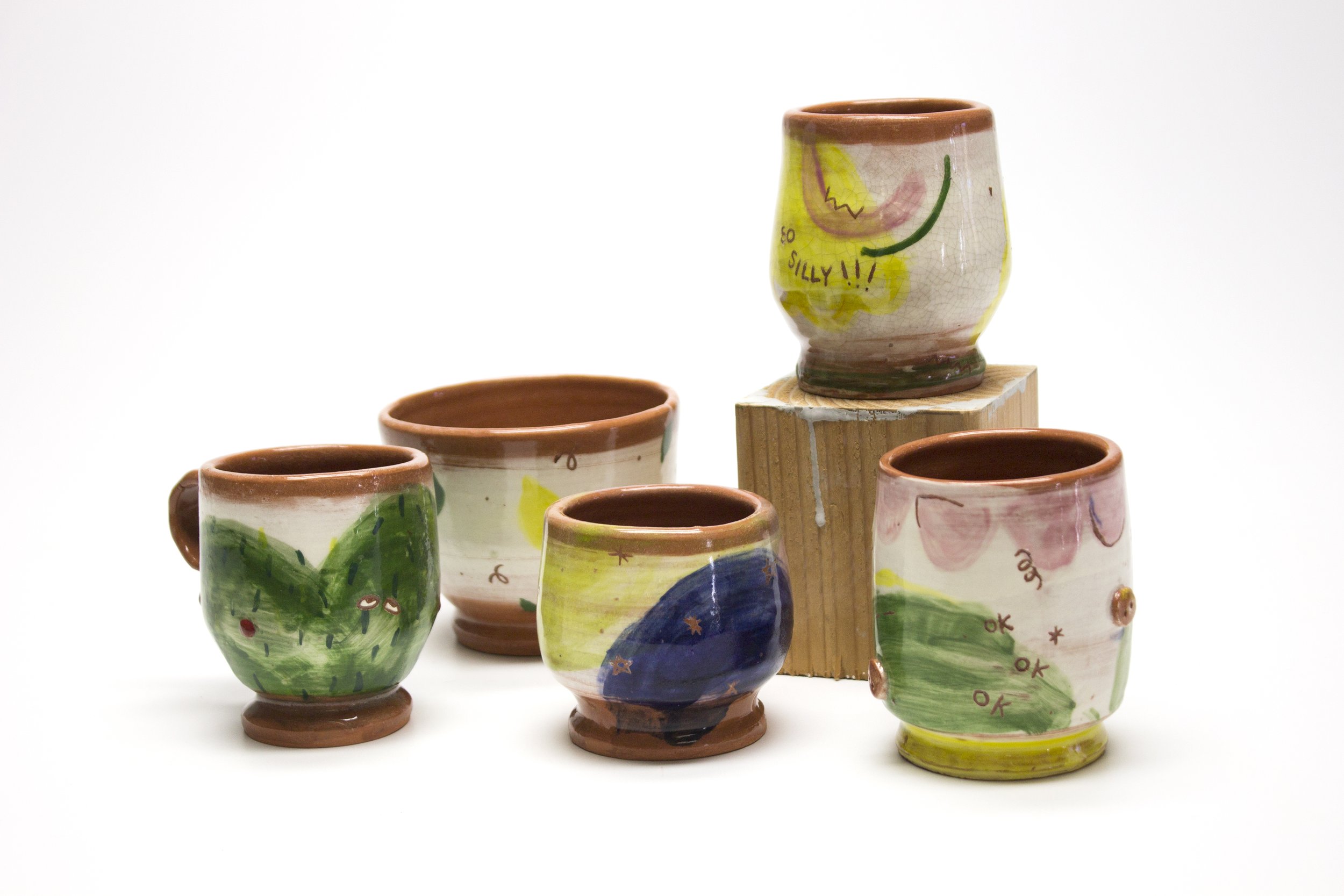 Five hand-painted ceramic mugs with colorful abstract designs, displayed on a white background with one mug elevated on a wooden block.