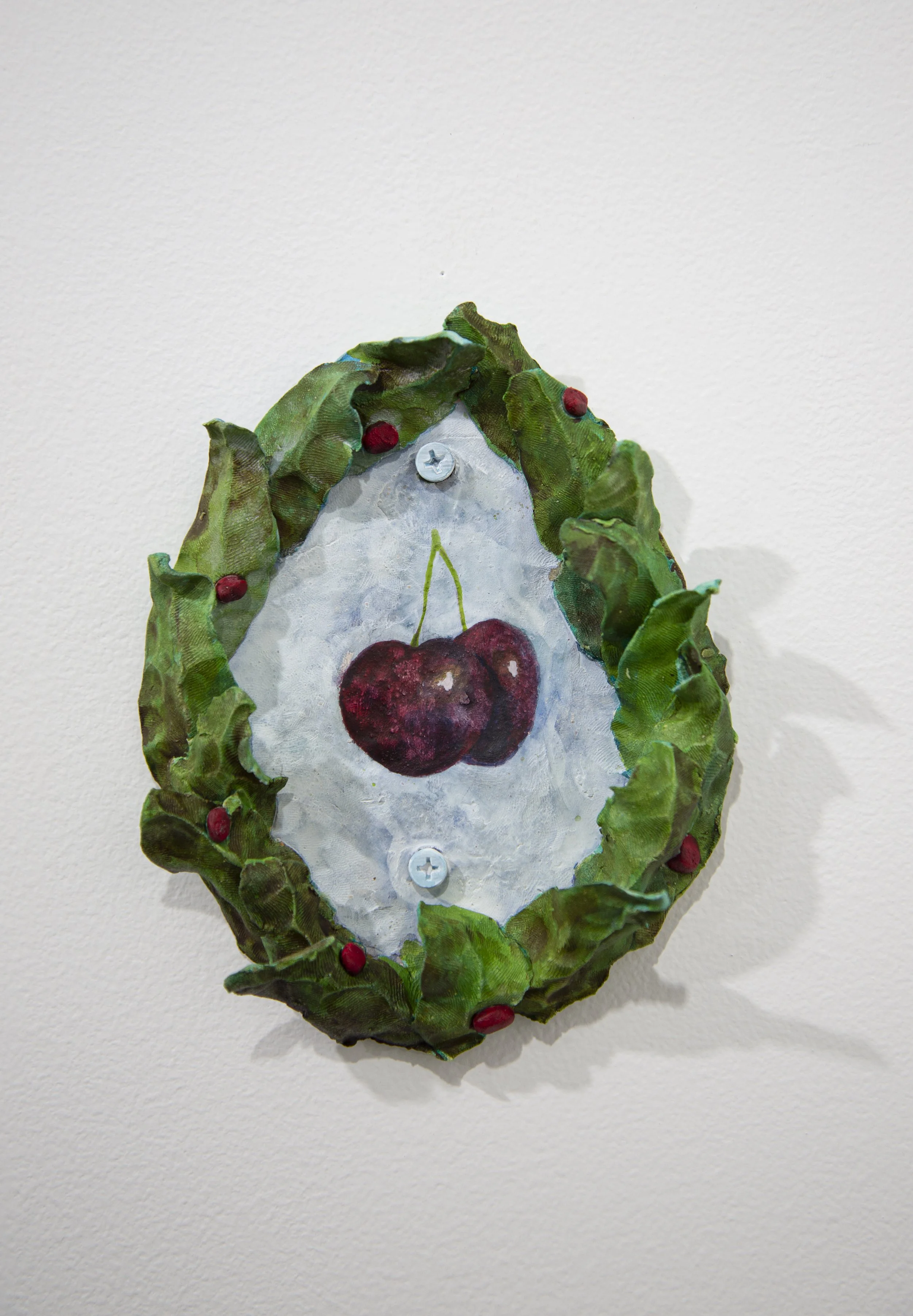 Mixed media artwork featuring a painted pair of cherries on a white background, framed with green textured leaves and red accents, attached to a wall with two screws.