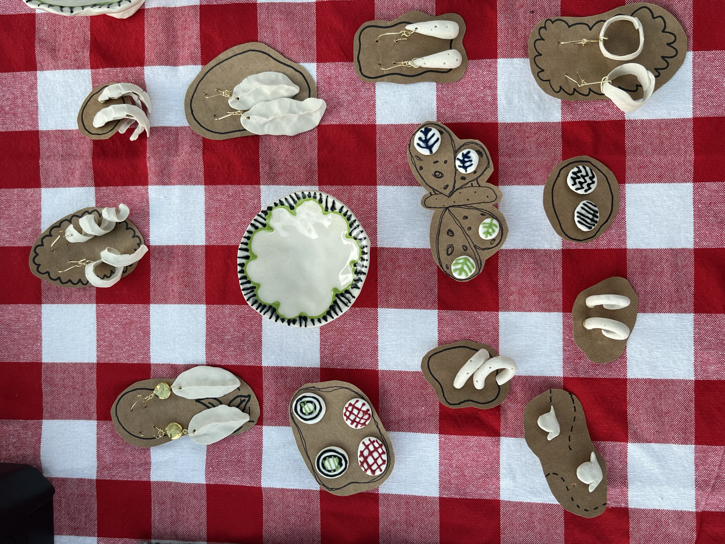 Display of handmade jewelry and decorative items on a red and white checkered tablecloth, including earrings, paper cutouts, and a decorated paper plate.