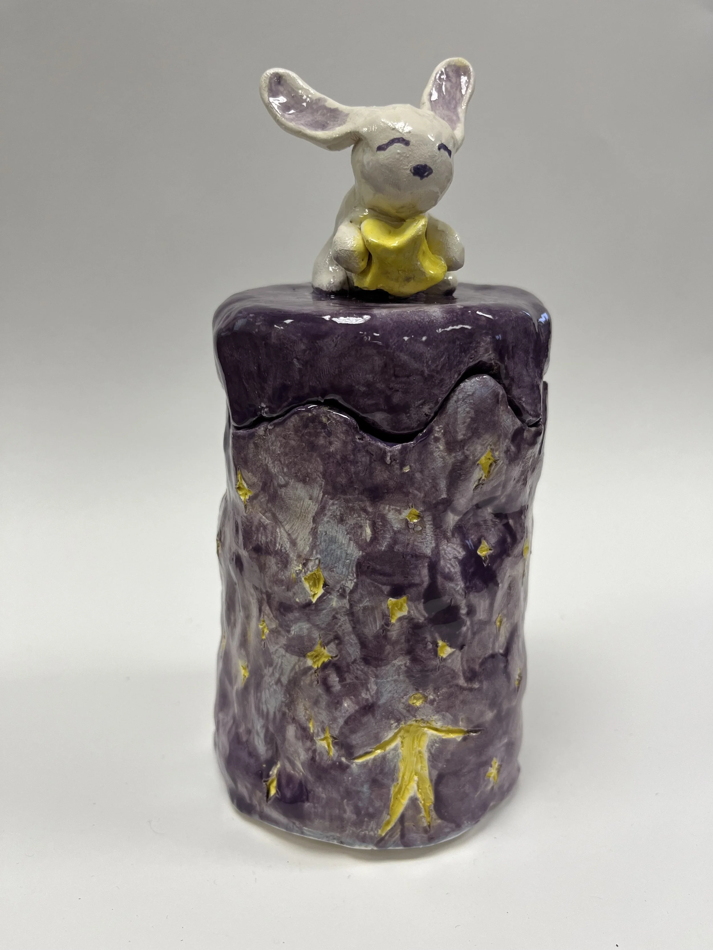 A small ceramic sculpture of a cute mouse with large ears, holding a yellow star, sitting on a purple cylindrical base decorated with small yellow stars and moon shapes.