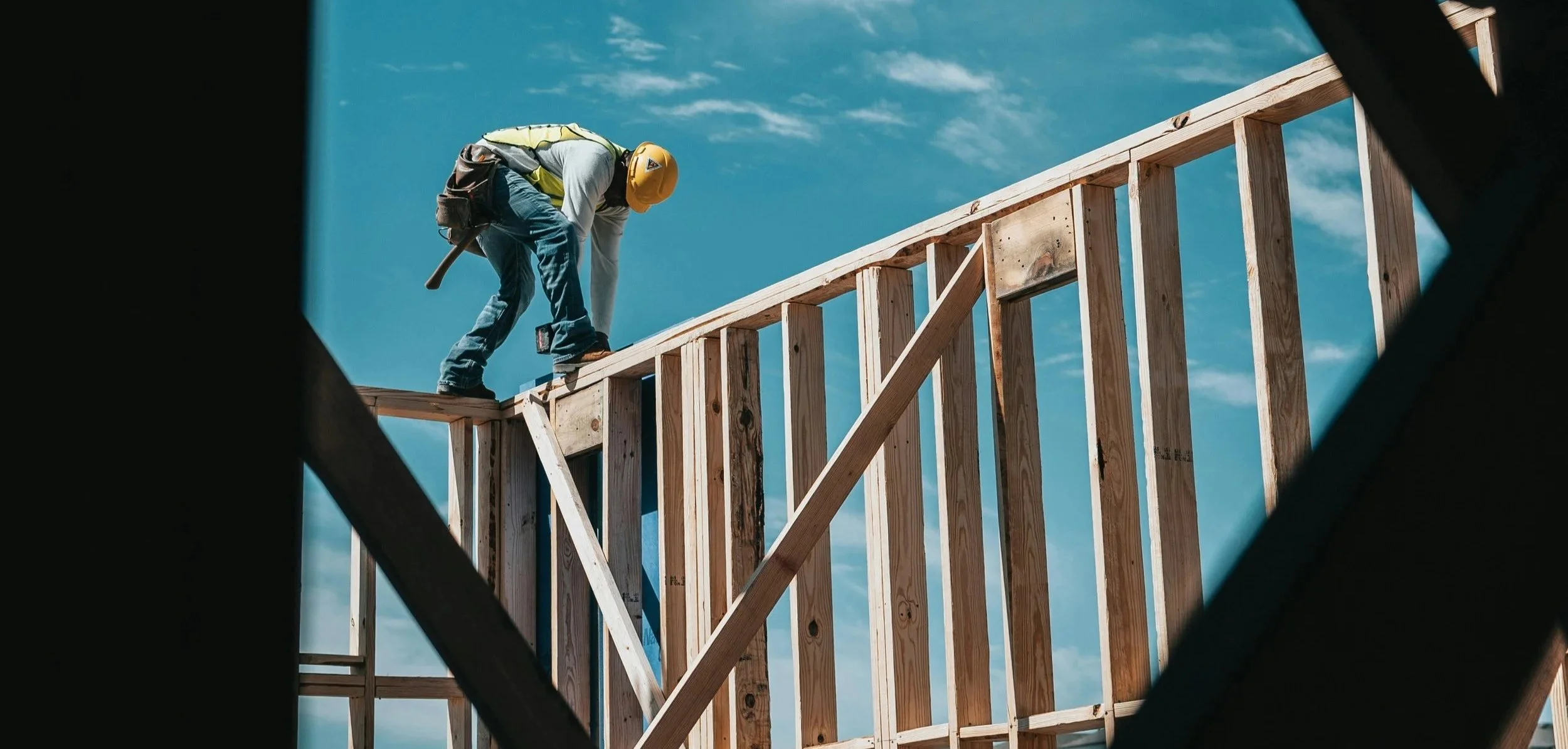 A construction worker wearing a hard hat standing on top of a wall of studs that they are building.