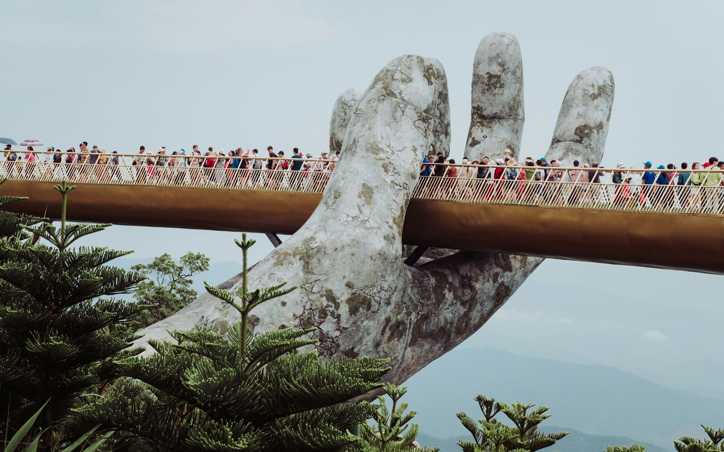 The Golden Bridge in Vietnam, featuring a giant hand-shaped support, is crowded with tourists walking across the pedestrian span, with lush green trees in the foreground and mountains in the background.