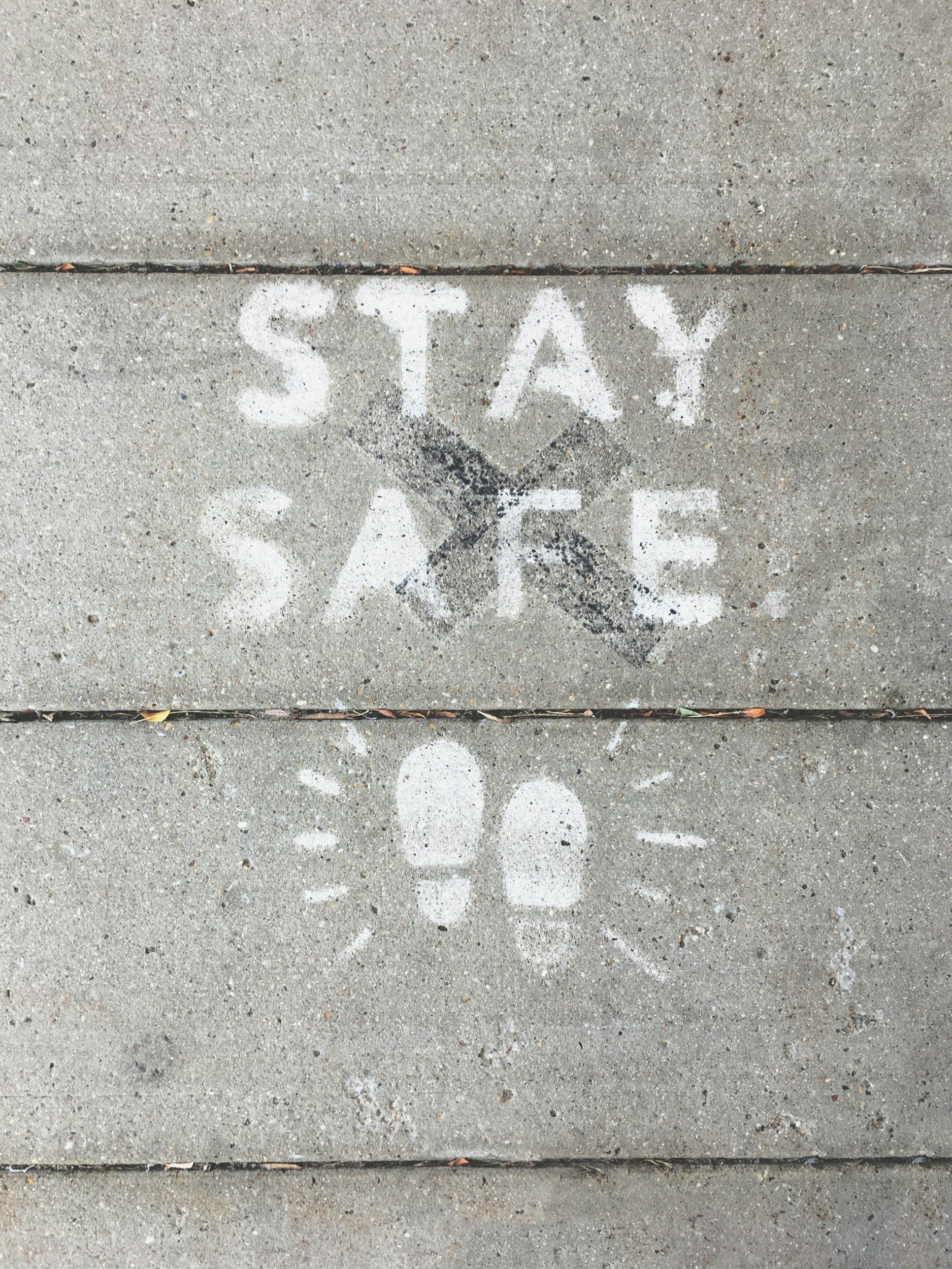 White sidewalk chalk markings with the words 'STAY SAFE' above a black X, and a white footprint with radiating lines below, on a concrete sidewalk.