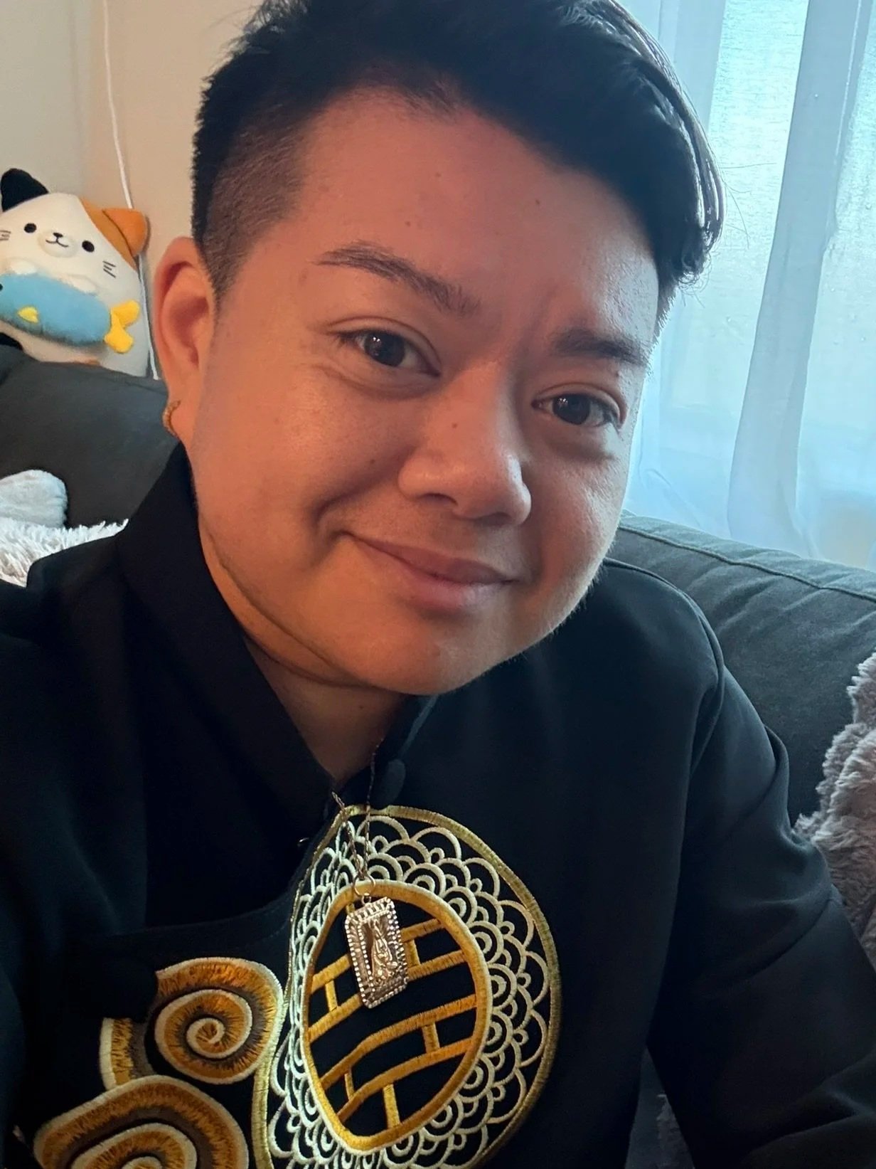 A person smiling, wearing a black garment with gold embroidery and jewelry, sitting on a sofa with a plush toy in the background, in a room with light coming through a window.