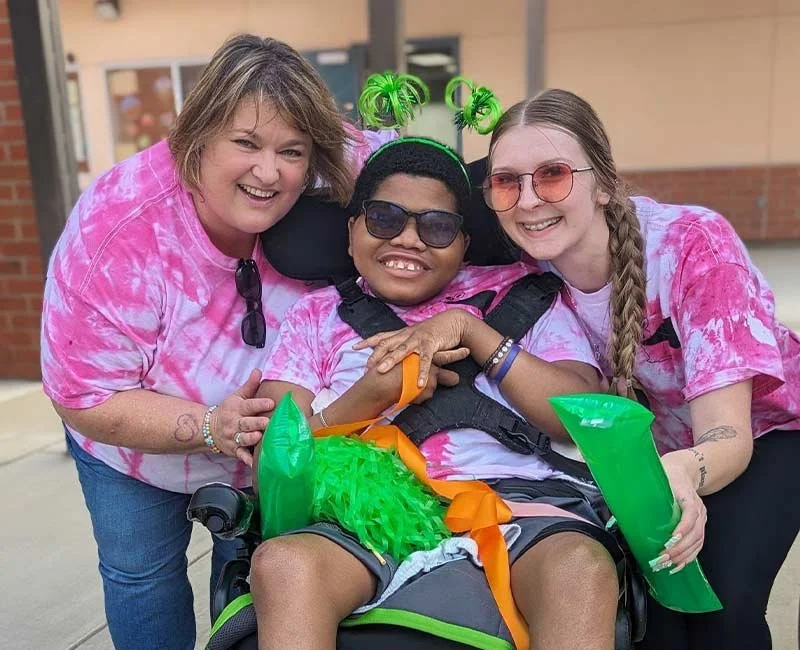 Three women smiling outdoors, one in a wheelchair with an inflatable dragon tail, all wearing pink tie-dye shirts.