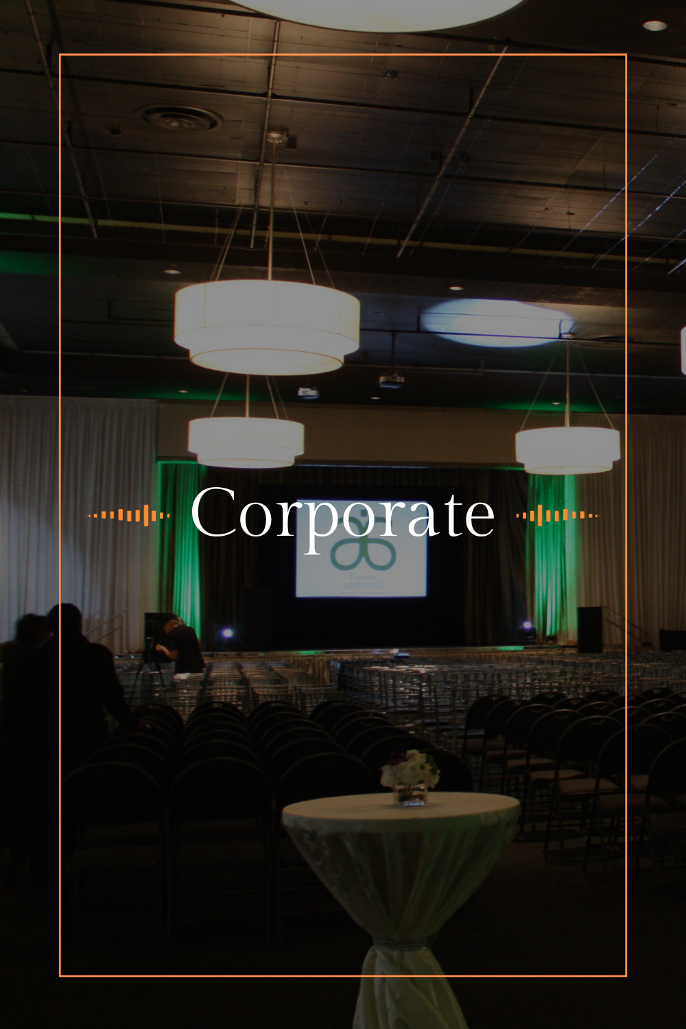 An empty conference hall with rows of chairs, a stage with a large screen displaying a logo, and hanging ceiling lights.