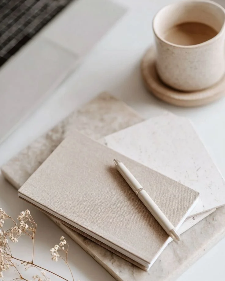 A beige notebook with a white pen resting on top, placed on marble and paper surfaces. In the background, there is a white cup filled with coffee on a matching saucer.