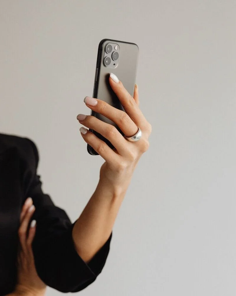 A person's hand holding a smartphone with a triple-camera setup, with a plain background and part of their arm in a black sleeve visible.