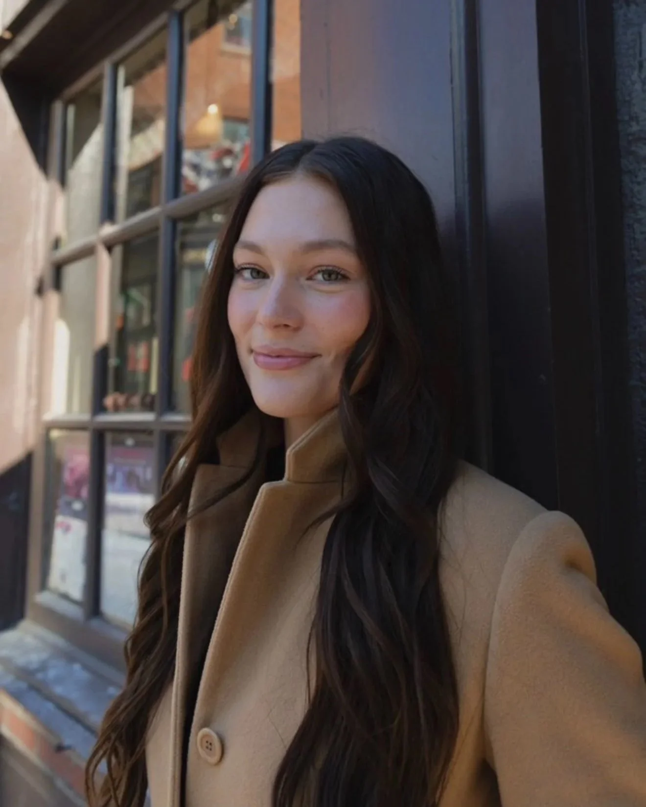 A young woman with long wavy dark hair and a light tan coat standing outside near a black wooden wall and large window with a brick building in the background.