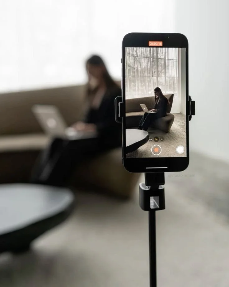 A woman sitting on a sofa using a laptop, seen through a smartphone mounted on a tripod recording or live streaming her.