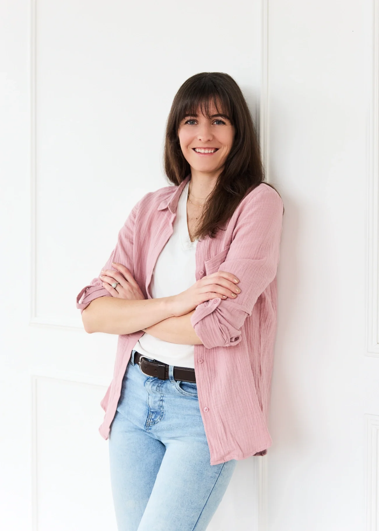 Cassie Shelley wearing a pink shirt and light blue jeans, standing against a white wall, smiling with arms crossed.