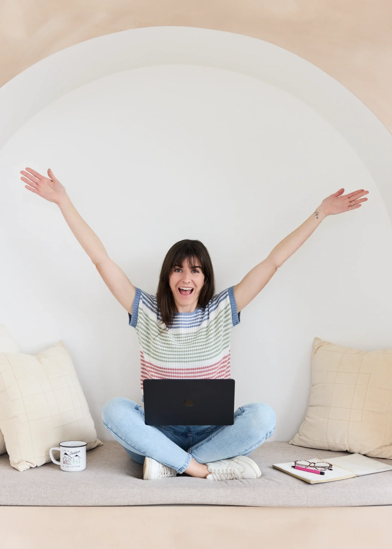 Cassie Shelley sitting cross-legged on a beige couch with a laptop on her lap, smiling with her arms raised.