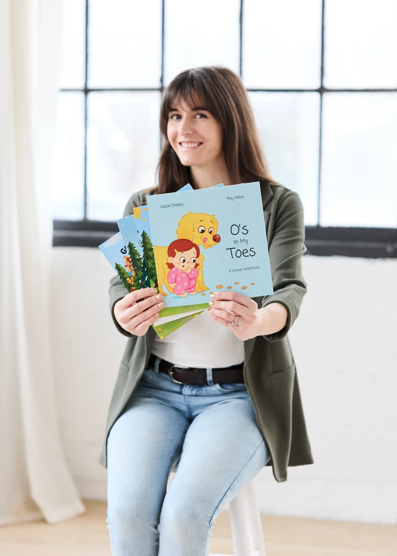 Cassie Shelley sitting on a white stool, holding her children's books out in front of her.
