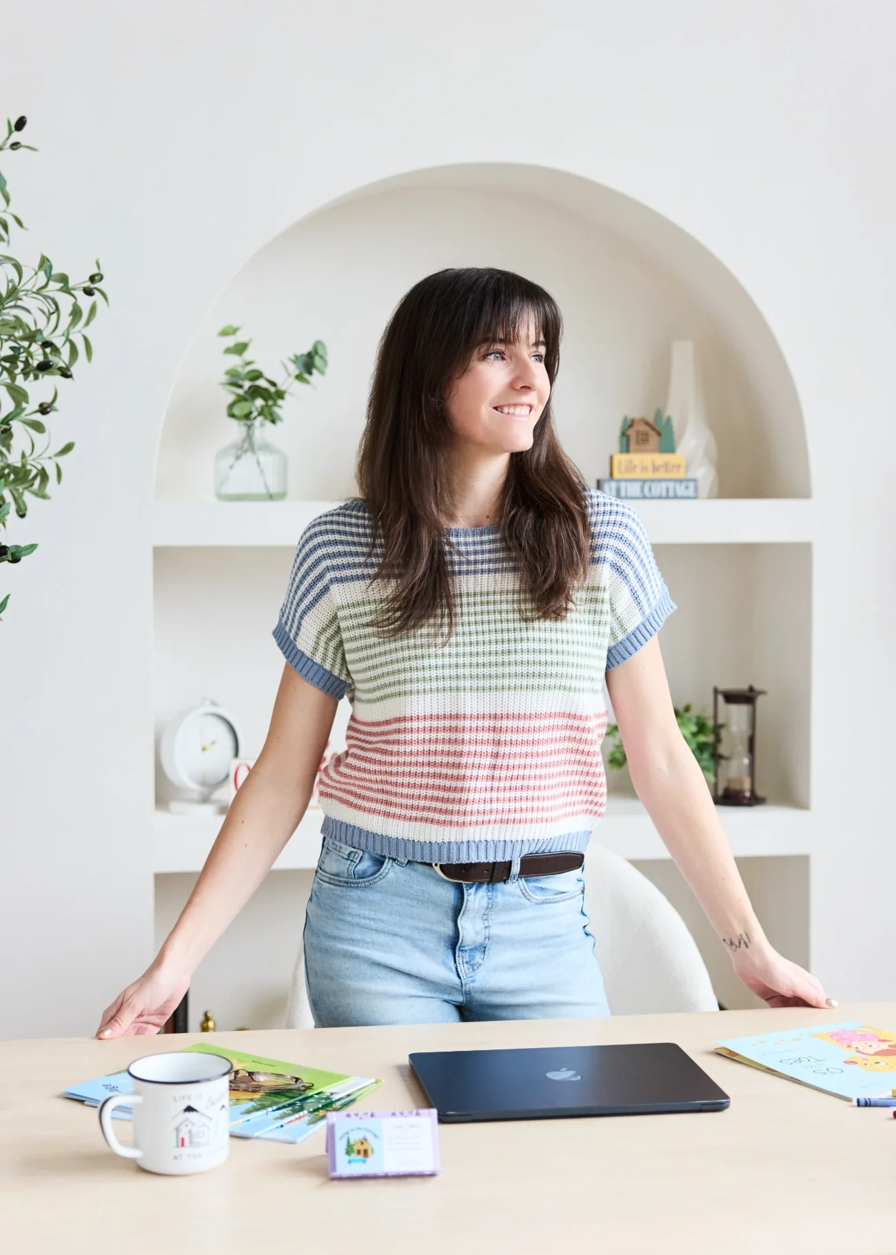 Cassie Shelley standing behind a desk, smiling and looking to her right. The desk has a closed laptop, a cup, magazines, and children's books.