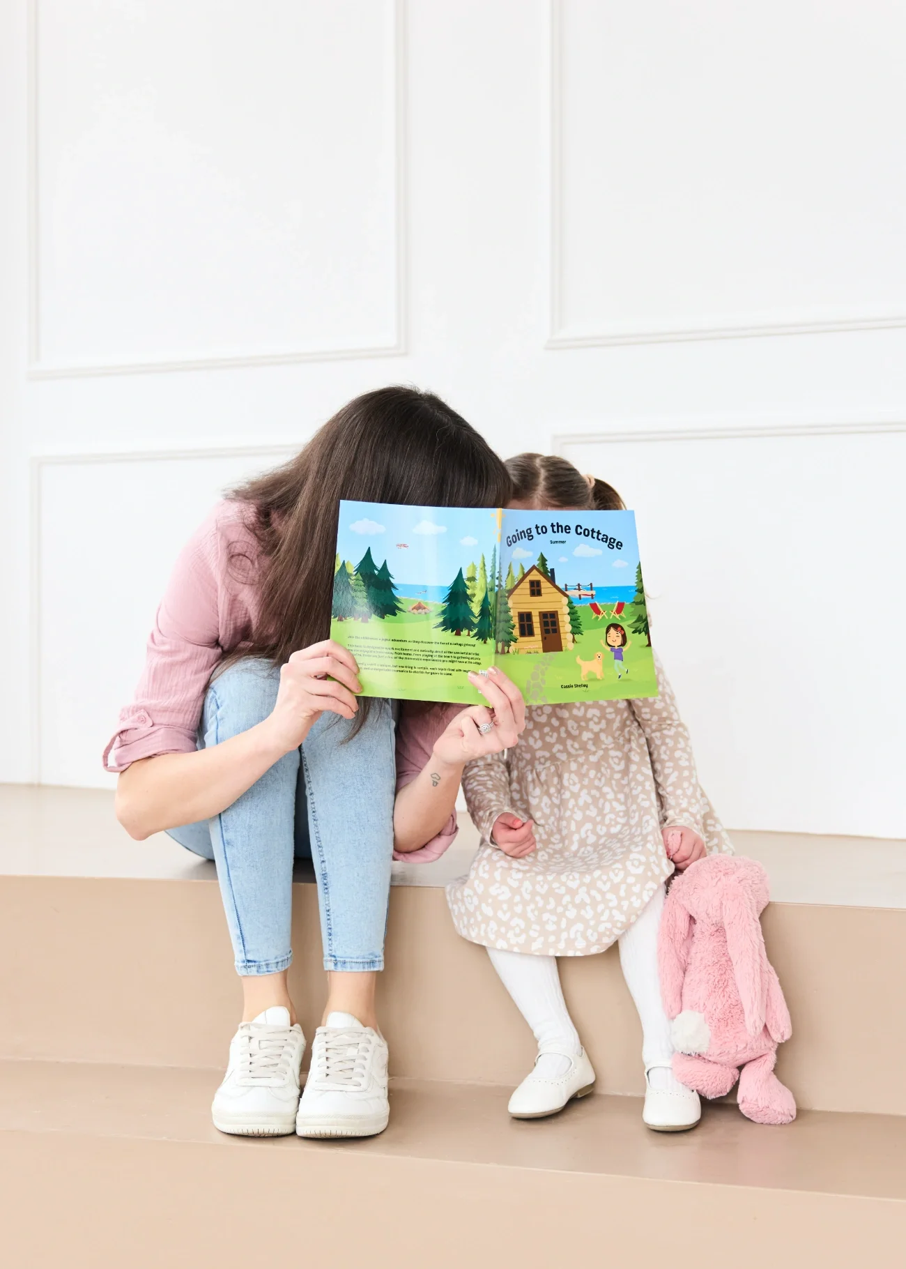 Cassie Shelley and her daughter sitting on a beige platform against a white wall reading 'Going to the Cottage,' partially obscuring her face, while the girl holds a pink stuffed rabbit and a pink blanket.
