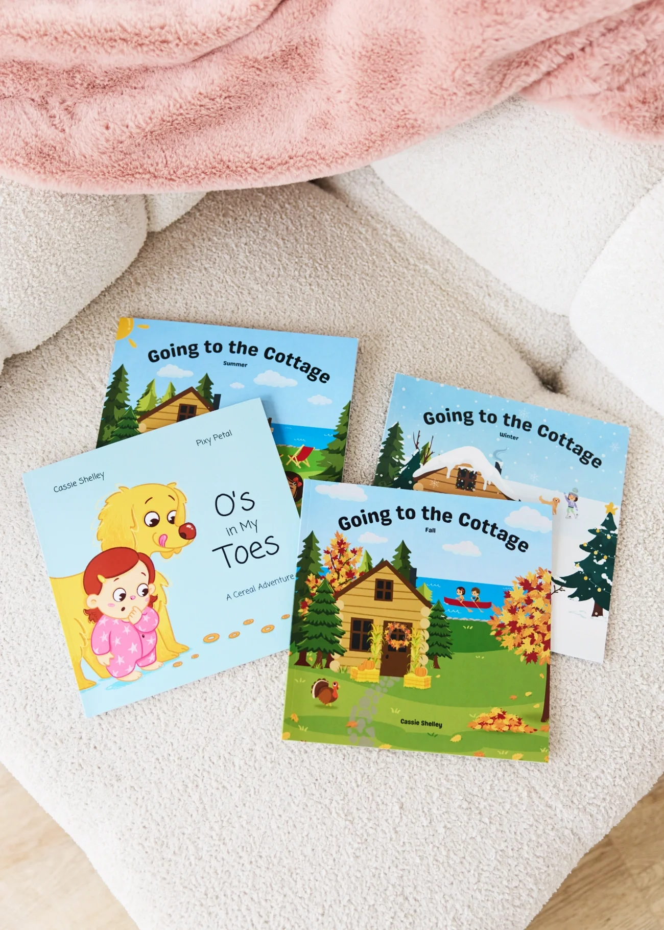 Four Cassie Shelley children's books titled "Going to the Cottage" Fall, Summer, and Winter, and "O's in My Toes: A Cereal Adventure" sitting on a light-coloured couch.