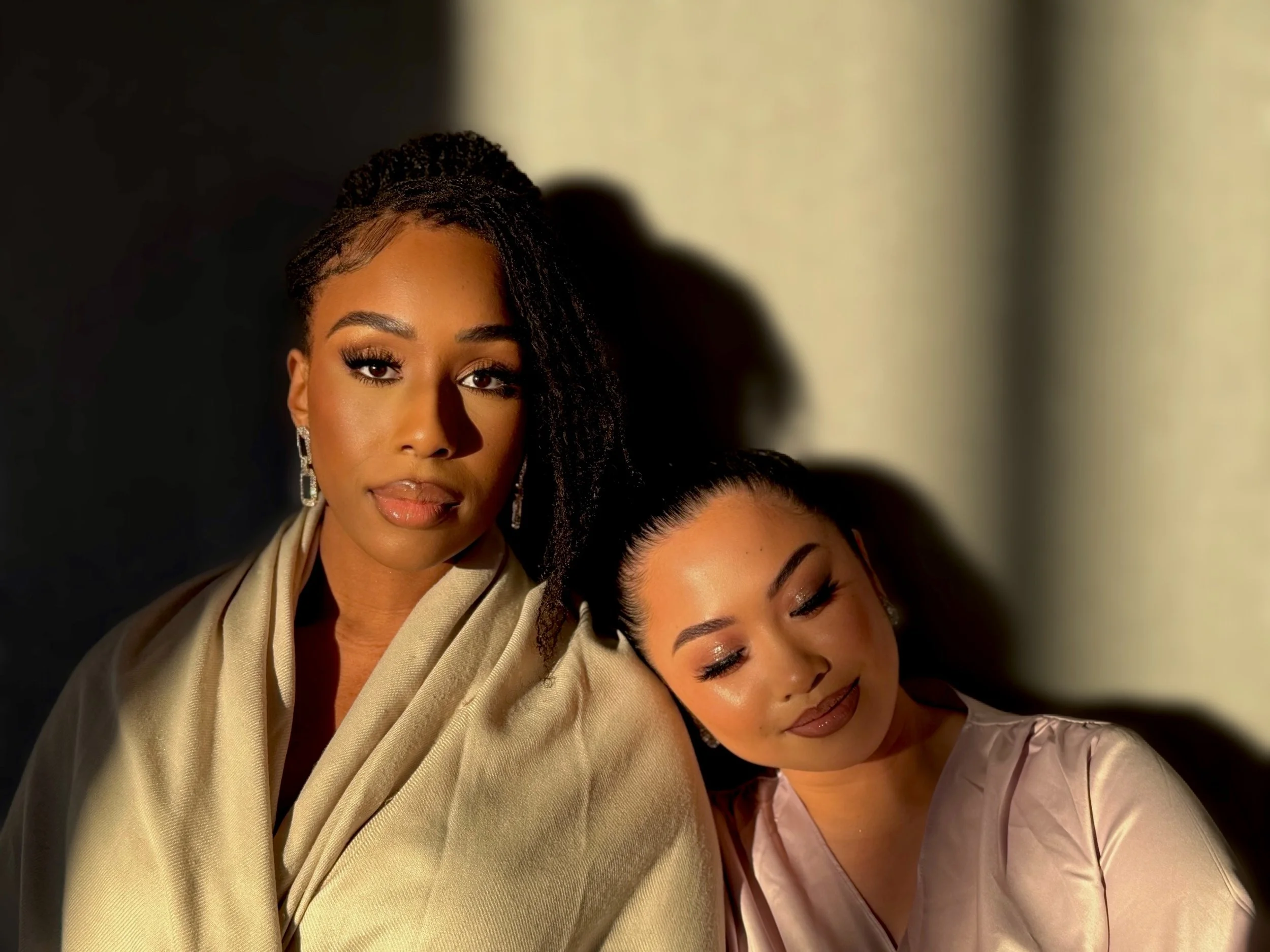 Two women, one with dark skin and braids, and the other with lighter skin and dark hair in a bun, pose closely together against a shadowed background, both dressed elegantly.