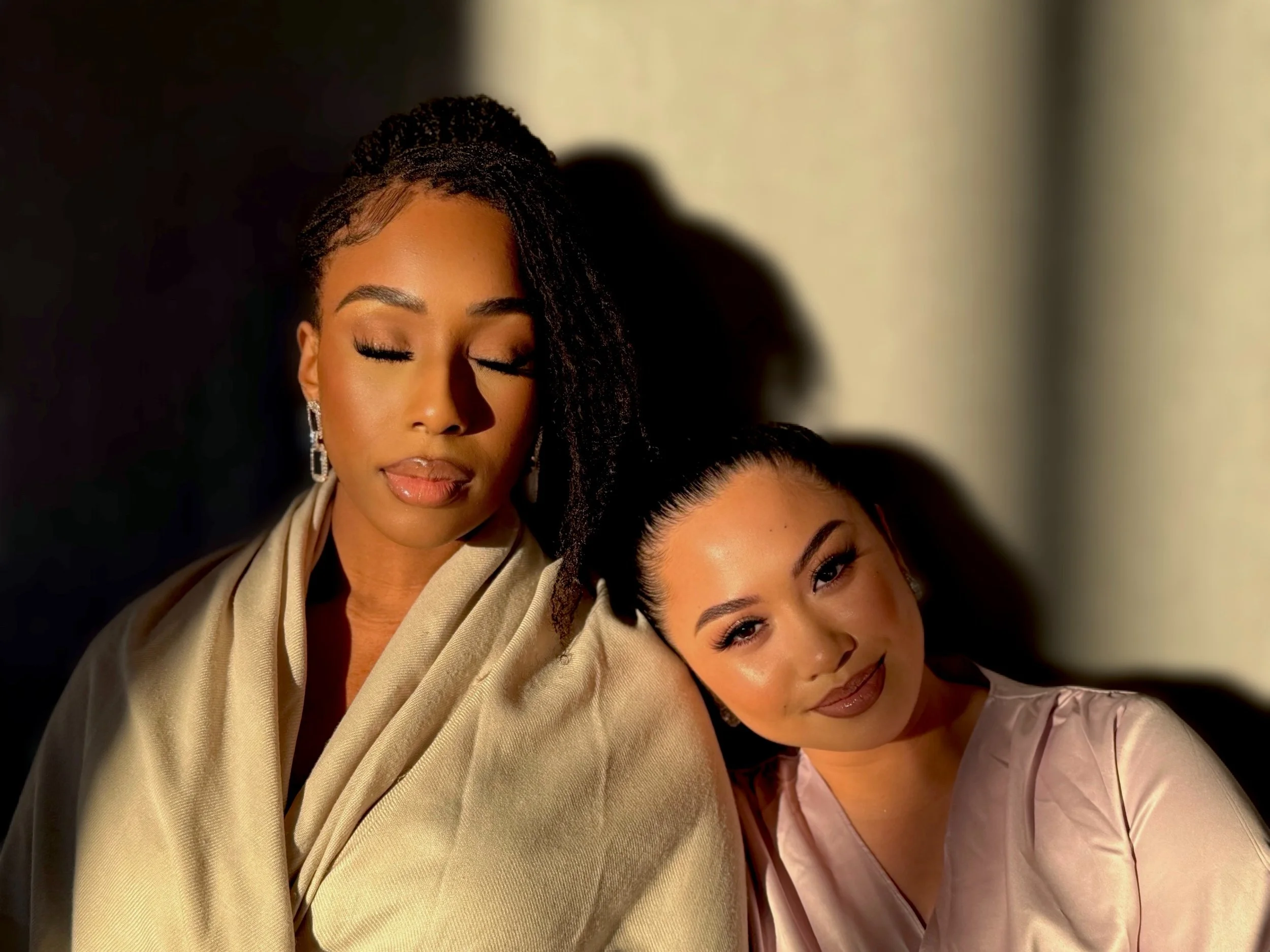Two women with their eyes closed, leaning on each other, with soft lighting.