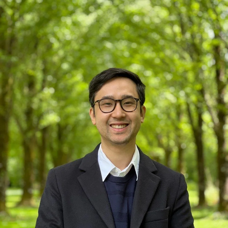 Portrait of a man with glasses smiling outdoors in a wooded park.