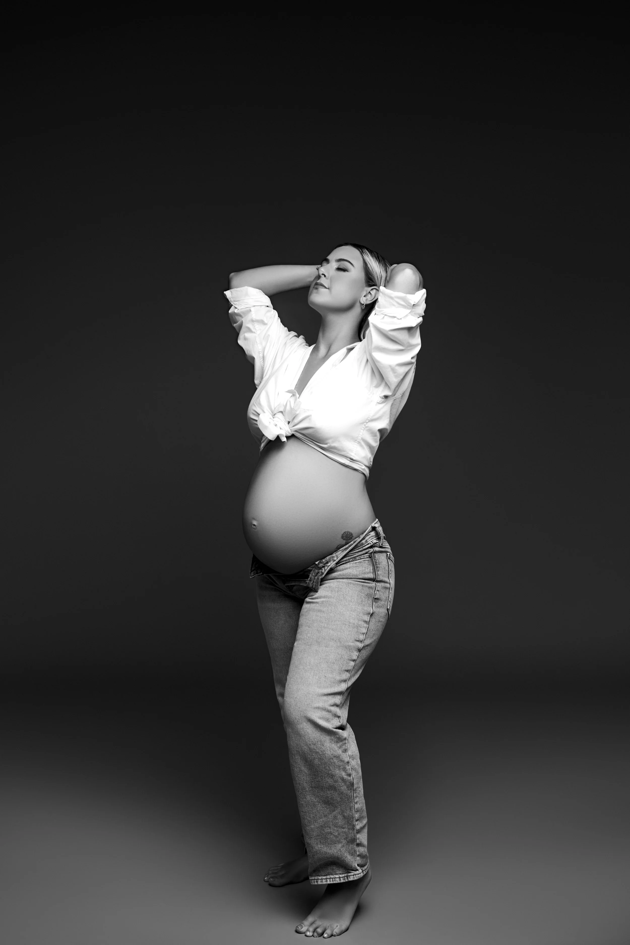Black and white photograph of a pregnant woman with arms raised and eyes closed, wearing a tied-up white shirt and jeans, barefoot, posing against a dark background.