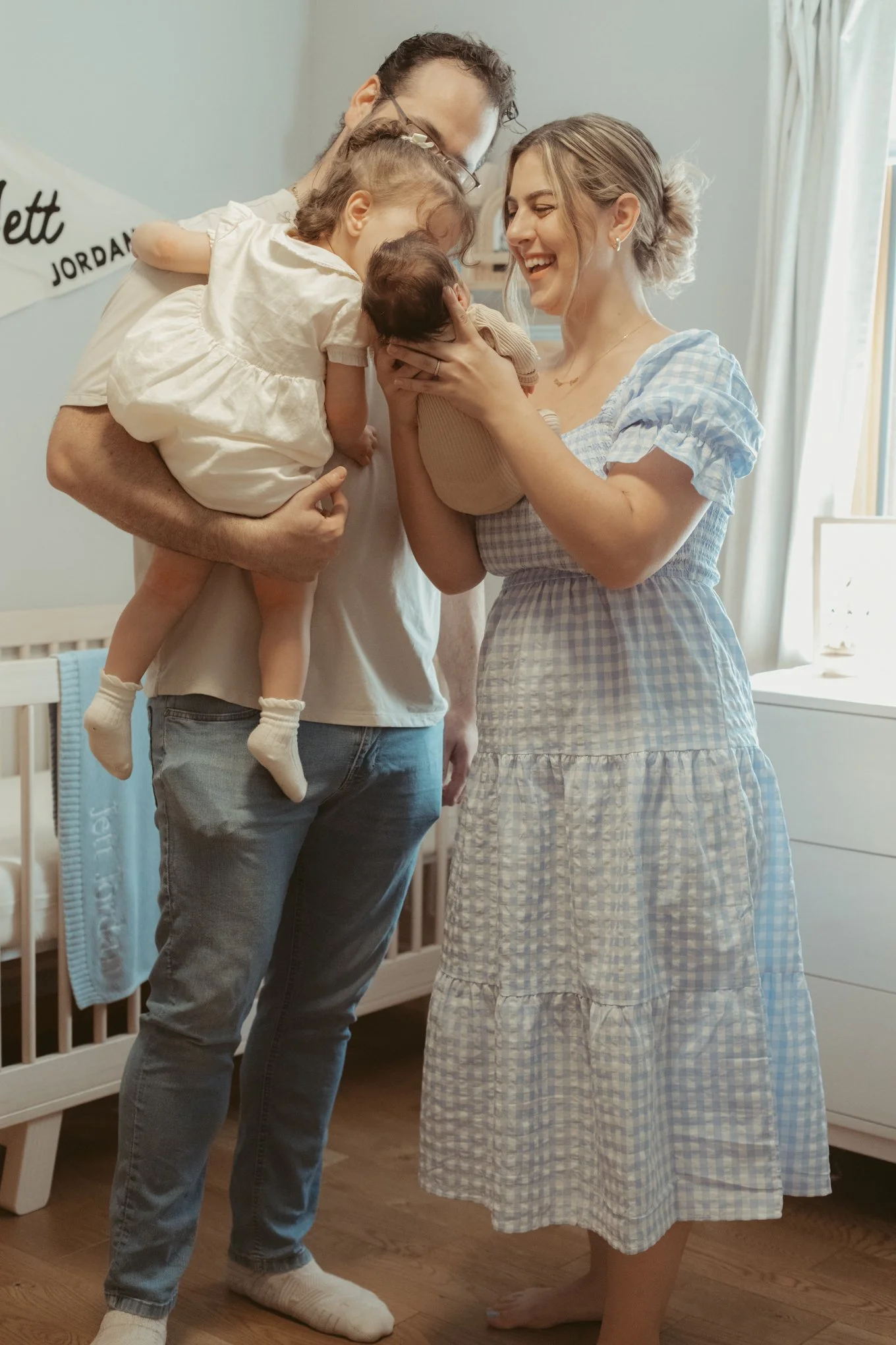 A family in a nursery, with a young couple holding their two children, one girl and one baby, as the woman admires the baby. Everyone is smiling and bonding closely.