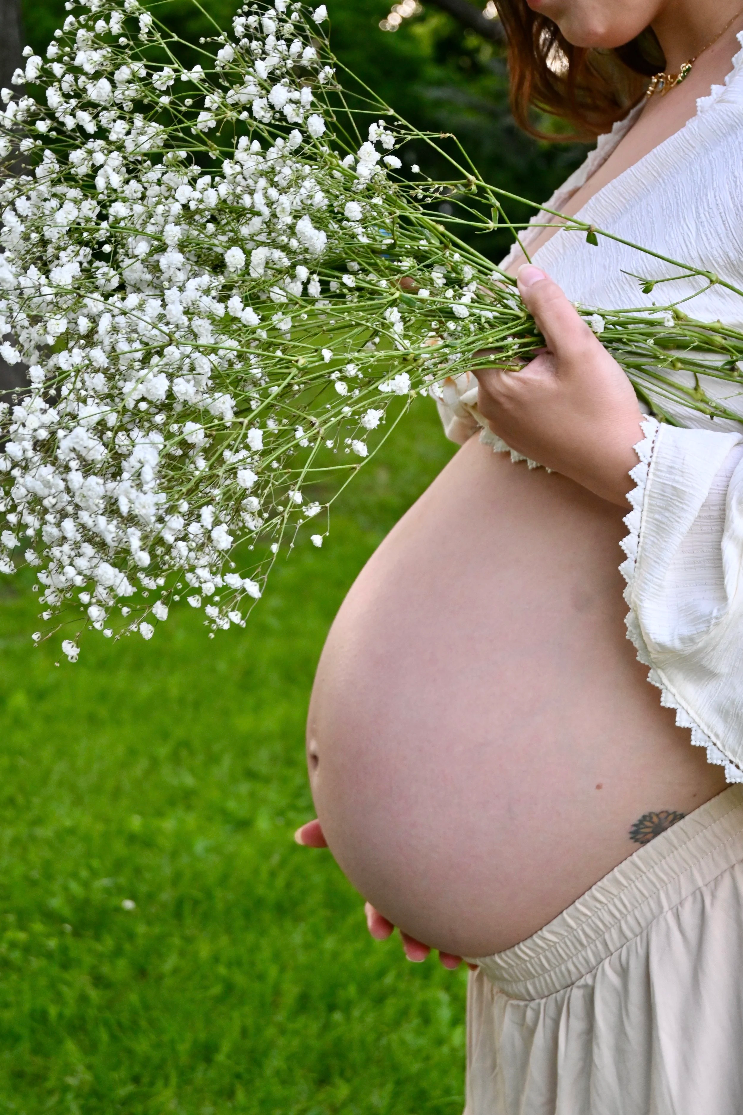 A pregnant woman wearing a white blouse and beige skirt holding a bouquet of white baby's breath flowers outdoors.