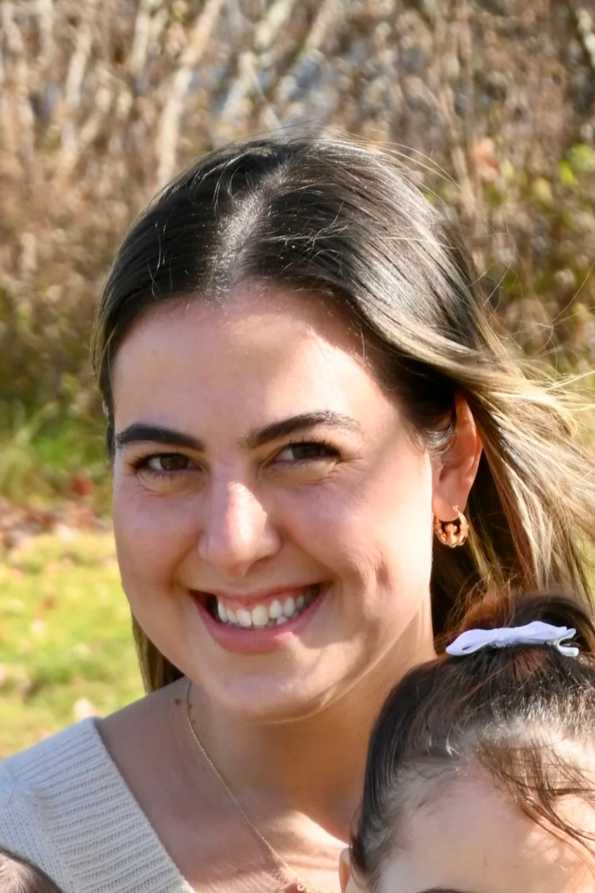 A woman smiling outdoors with greenery and trees in the background.