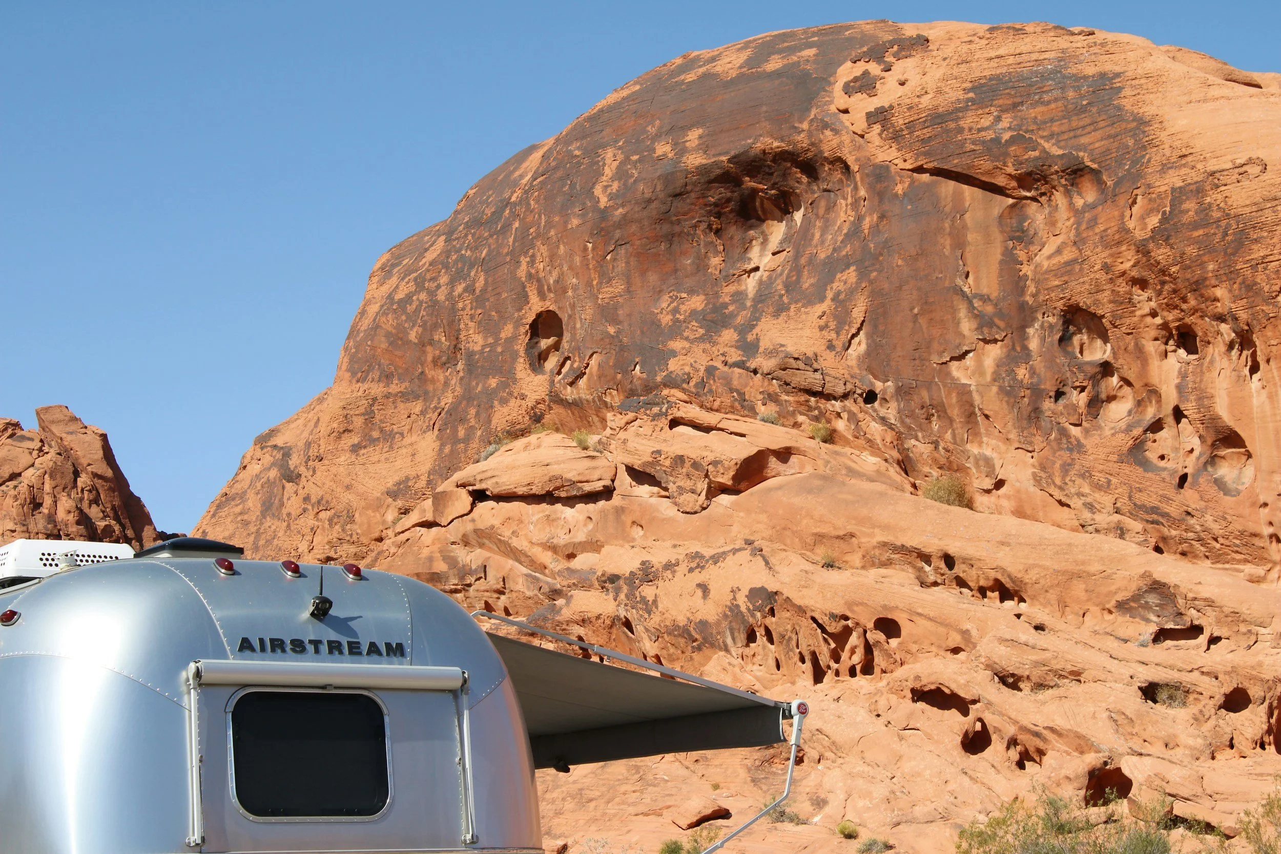 Silver Airstream camper parked near large red rock formations in a desert landscape under a clear blue sky.
