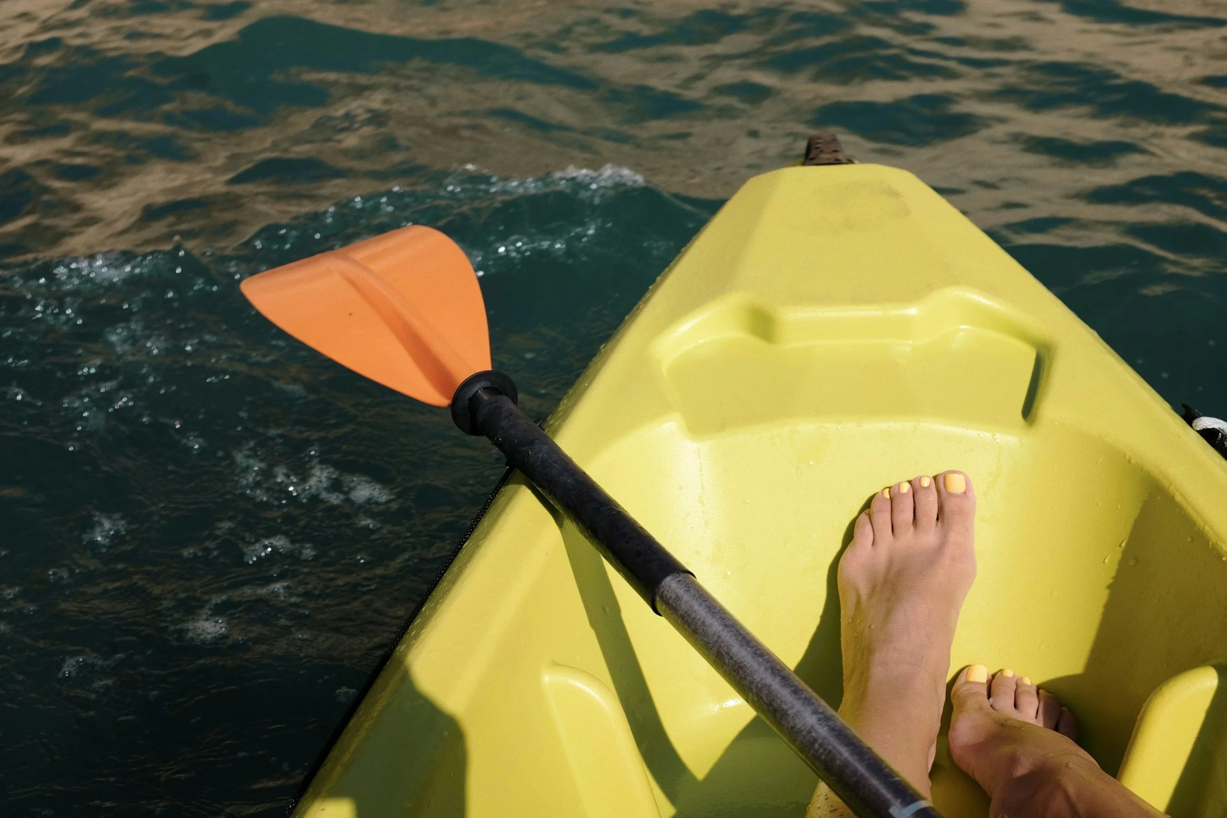 View from someone sitting in a yellow kayak with pedicure yellow toenails, paddling on the water with an orange paddle.
