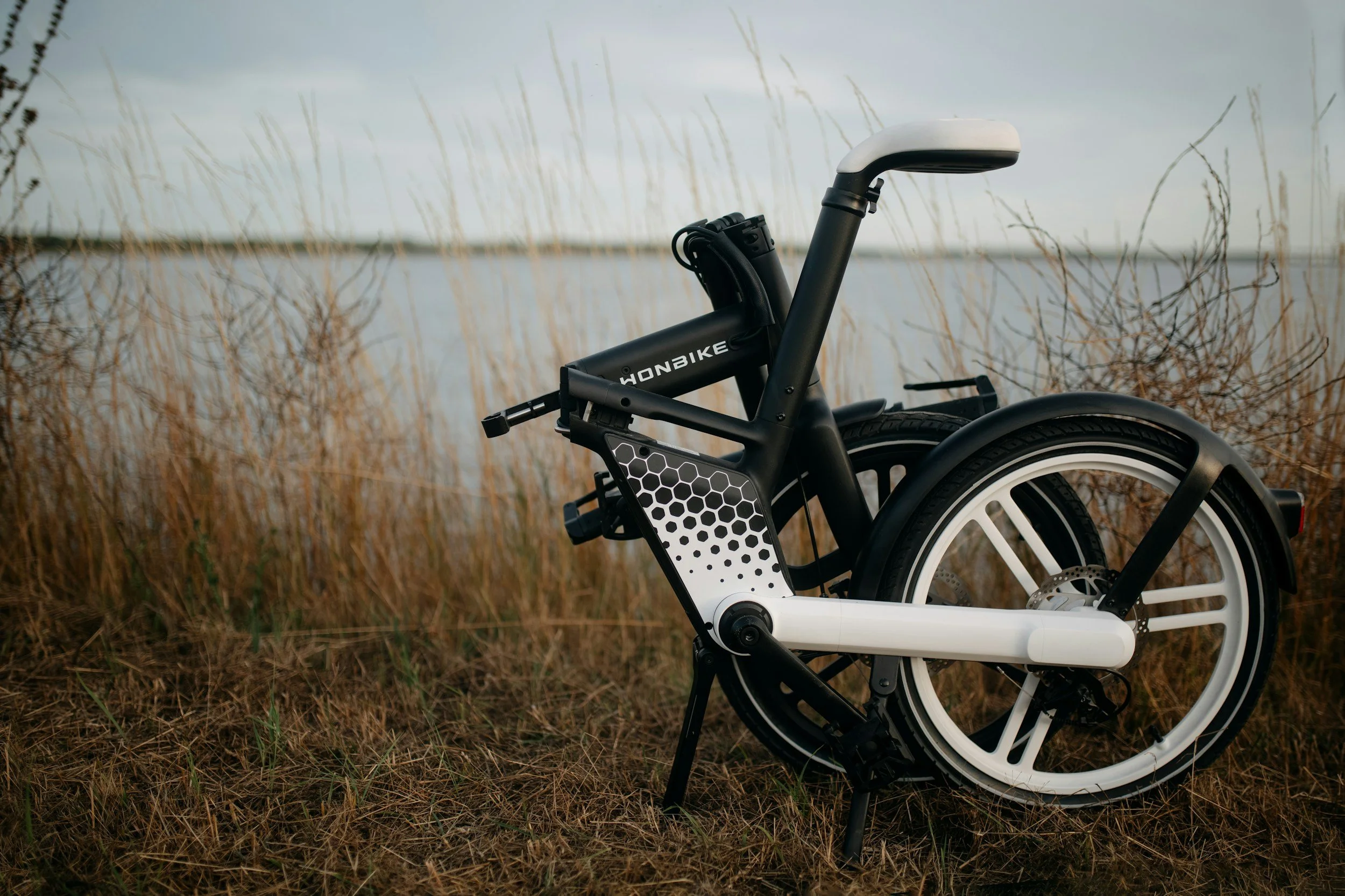 Folded black and white electric bicycle on grass near water with dried plants and cloudy sky in the background.