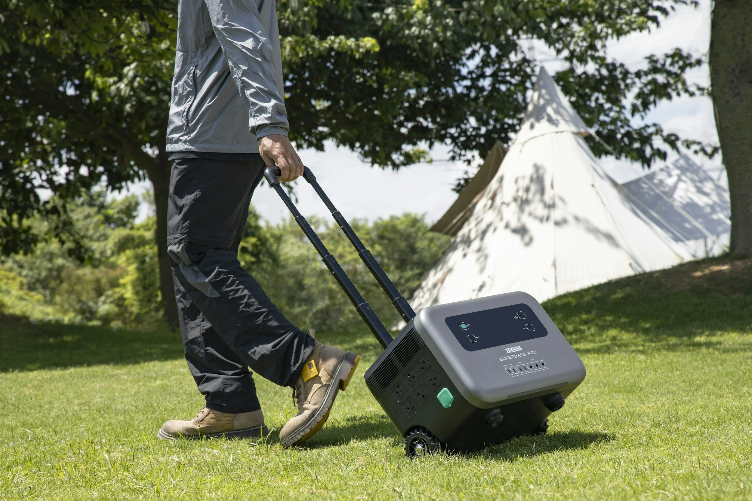 Person walking on grass in a park during daytime, pulling a portable power station on wheels, with trees and a white tent in the background.