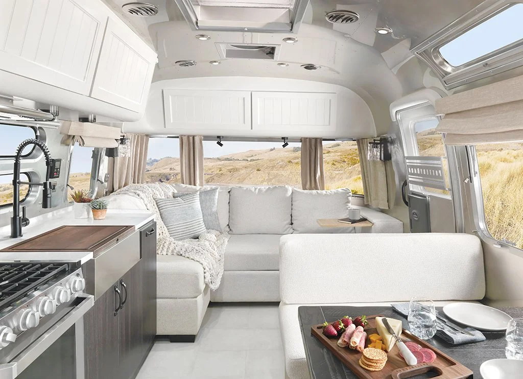Interior of an Airstream trailer with a cozy sofa, dining table with cheese and strawberries, kitchen area with stove and sink, and large windows showing a desert landscape outside.