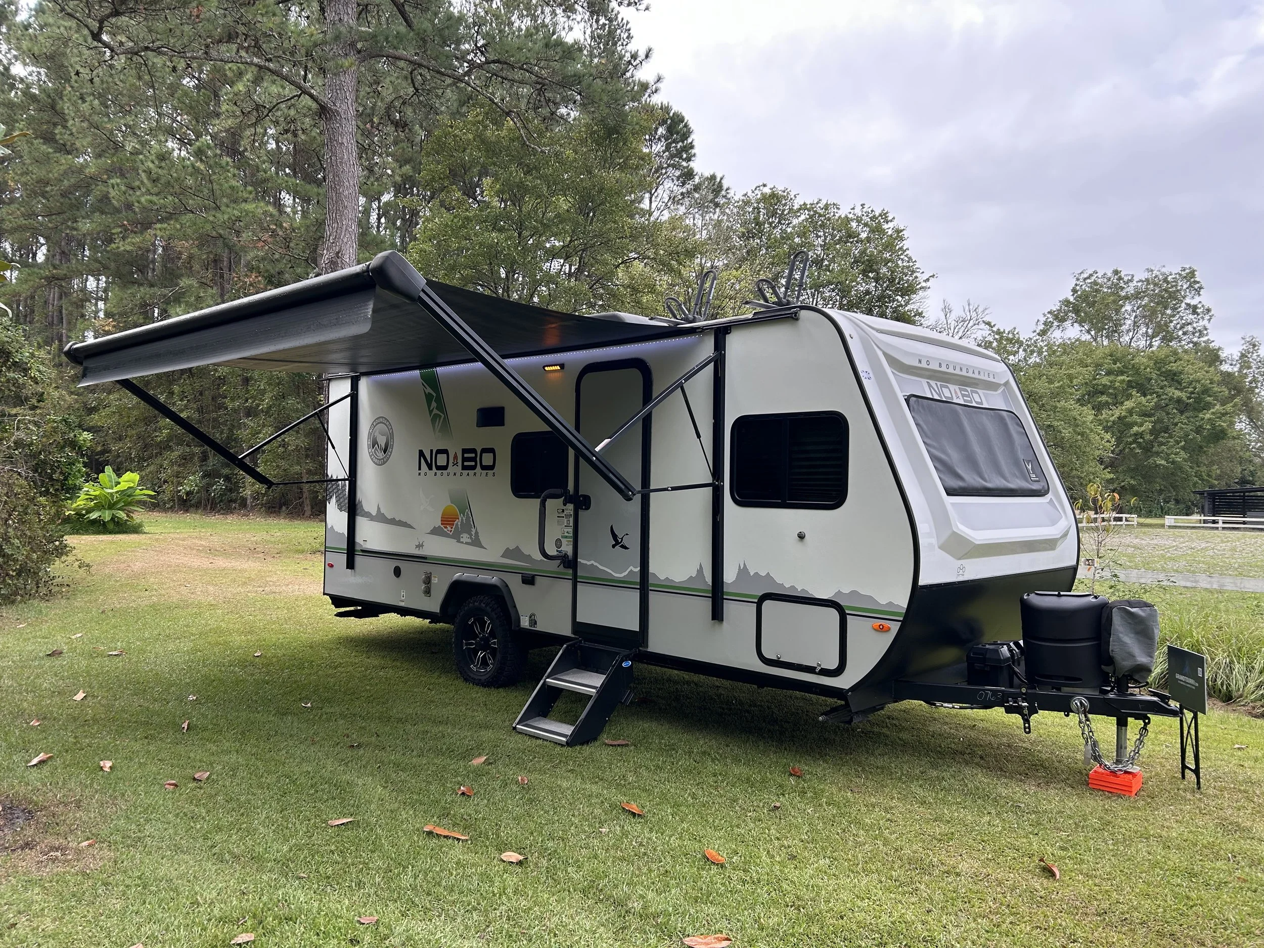 A modern travel trailer with an extended awning parked on a grassy area near trees with overcast sky.