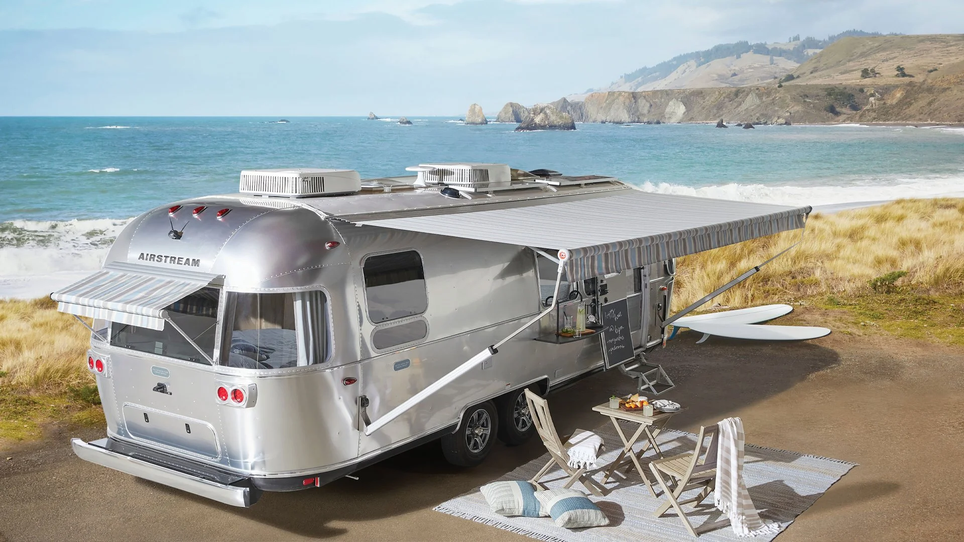 Aerial view of a silver Airstream camper trailer parked on a beach with an ocean and cliffs in the background. The camper has an awning, small outdoor table, chairs, and surfboards.