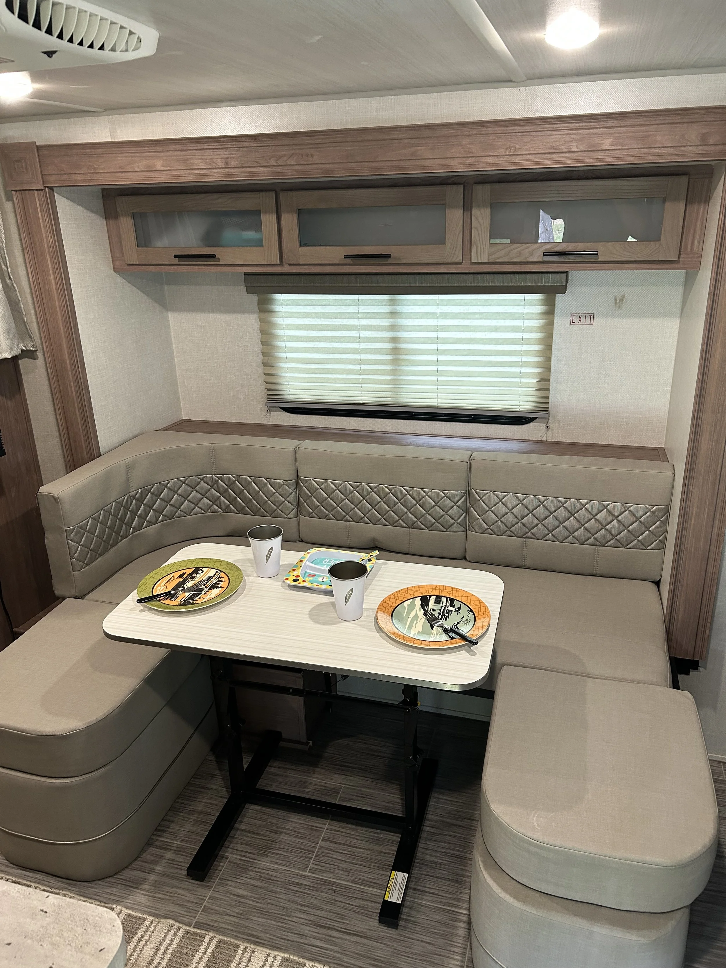 Inside an RV, a dining area with U-shaped beige cushioned seating, a white foldable table, two plates, two cups, and a children's tray on the table. There is a window with blinds and overhead cabinets.