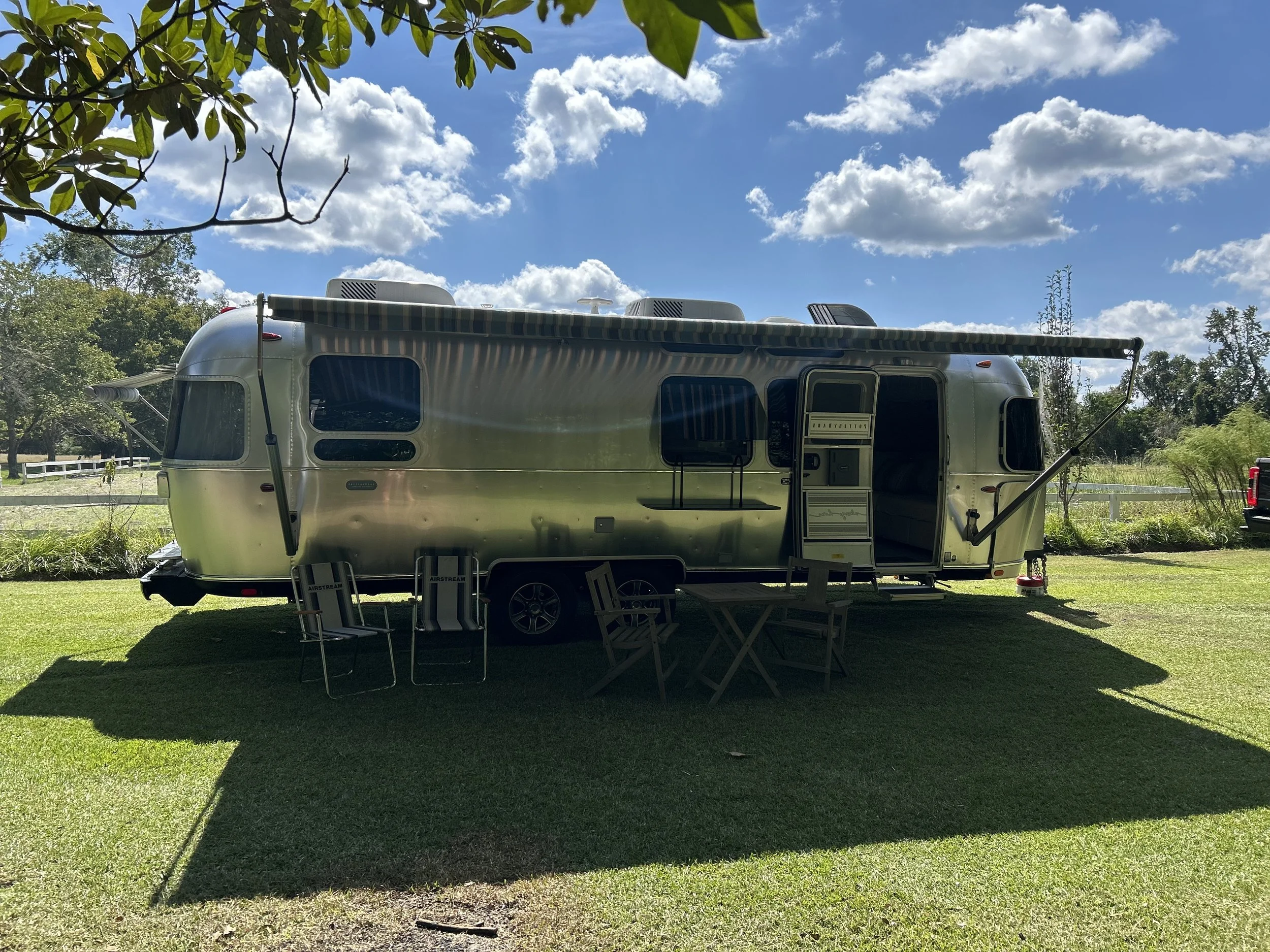 A shiny vintage Airstream travel trailer parked on a grassy area under a partly cloudy sky, with outdoor chairs and tables in front.